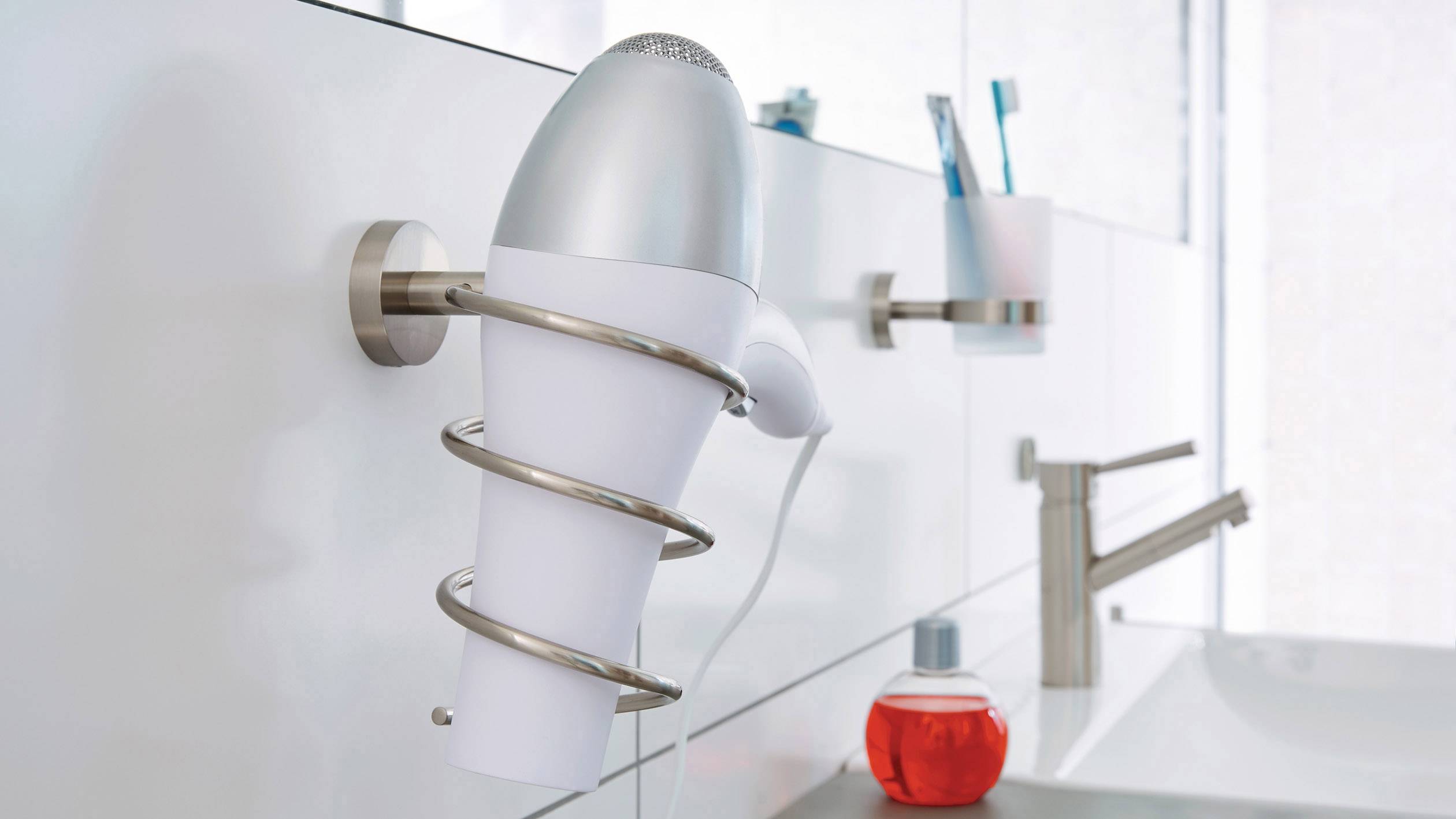 Ein Haartrockner hängt an der Wand im Badezimmer. Im Hintergrund sind Zahnbürsten und eine rote Flasche neben einem Wasserhahn.