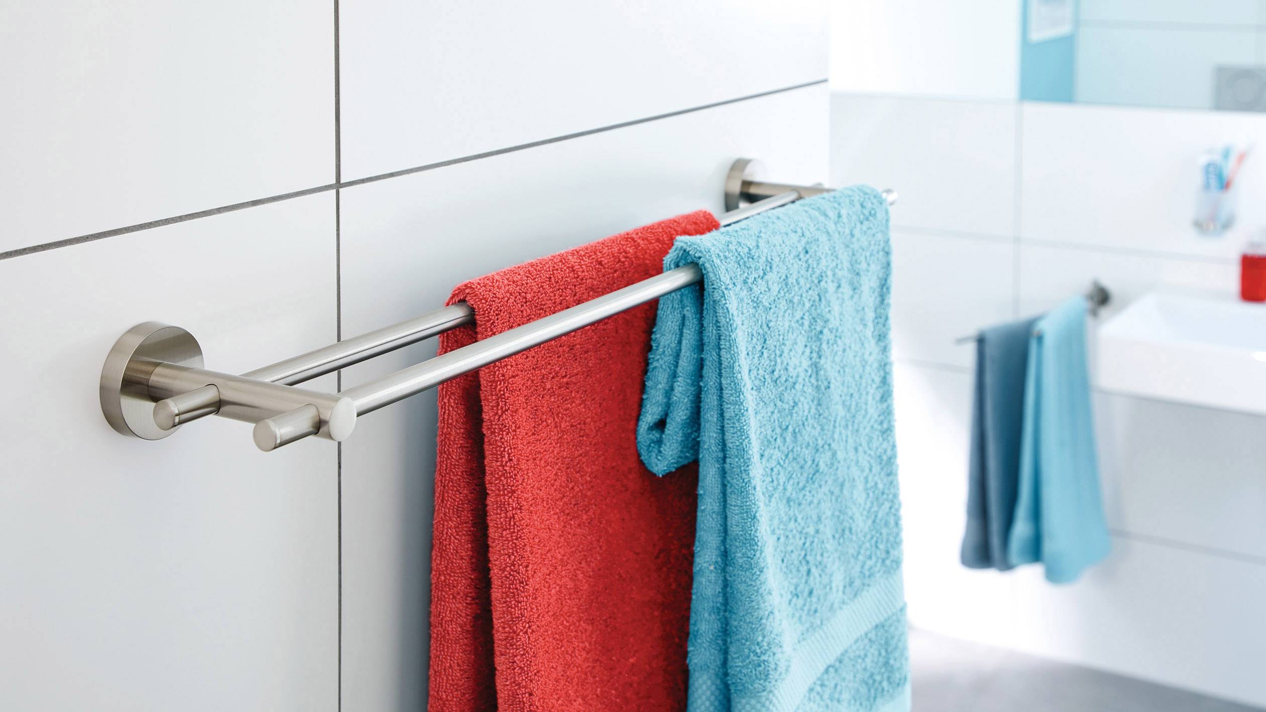 Ein Badezimmer mit weiß gefliesten Wänden. Ein Handtuchhalter trägt ein rotes und ein blaues Handtuch. Im Hintergrund ein Waschbecken.