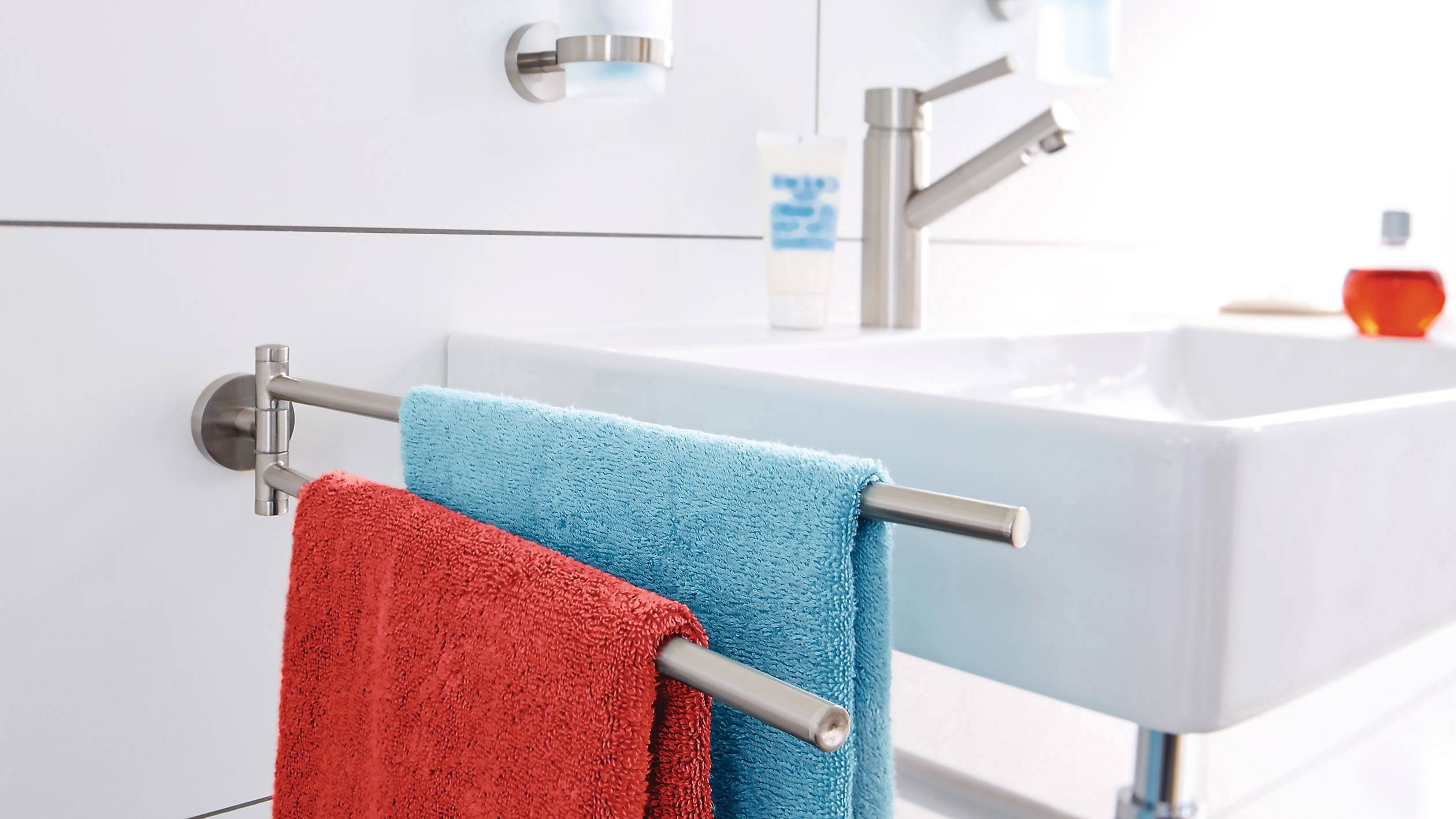 Ein Bad mit einem rechteckigen weißen Waschbecken, zwei Wandhandtuchhaltern mit roten und blauen Handtüchern, einem Wasserhahn und Pflegeprodukten.