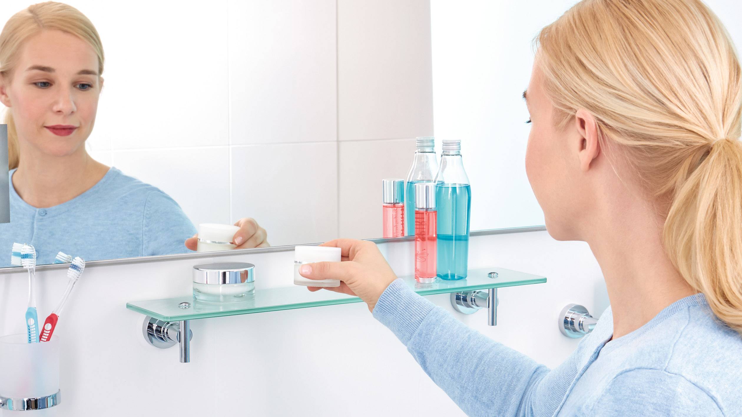 Eine Frau im Badezimmer nimmt eine Cremedose von einem Glasregal. Zahnbürsten und Mundwasser stehen daneben.