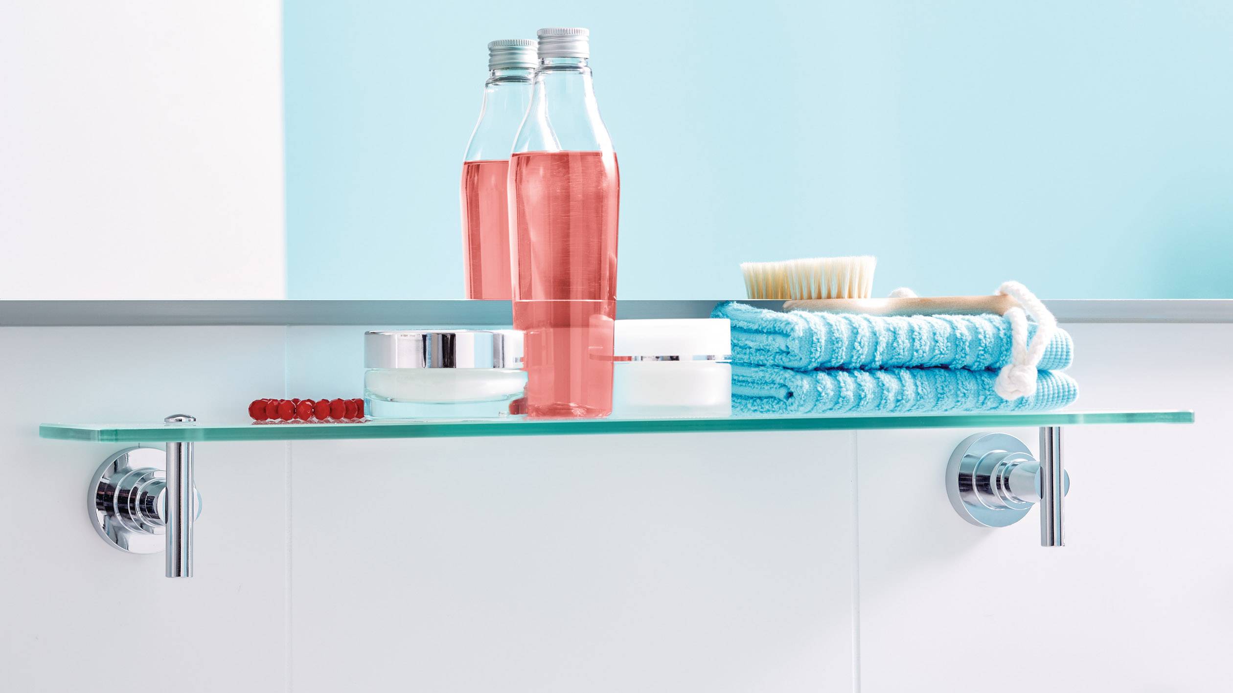 Pflegeprodukte, darunter zwei rosa Flaschen, ein Tiegel, ein Badetuch und eine Bürste, stehen auf einem Glasregal im Badezimmer.