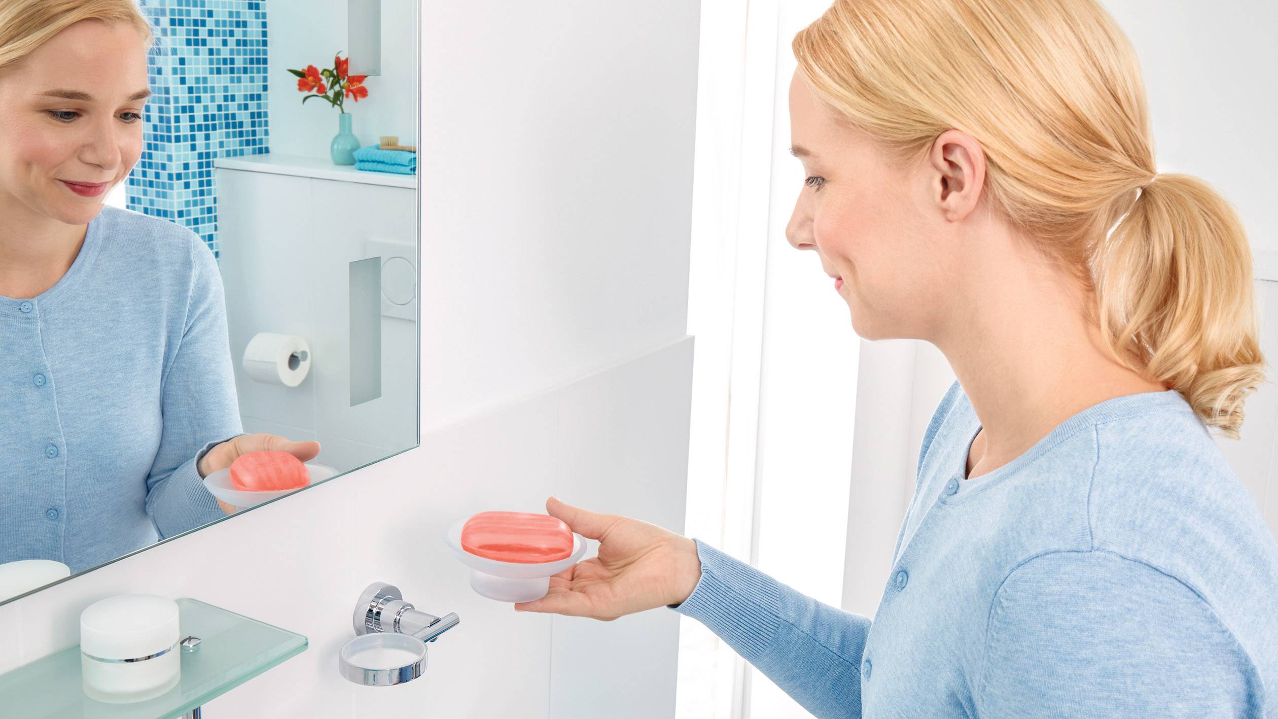 Eine Frau im Badezimmer hält eine Seifenschale in der linken Hand und schaut auf ihr Spiegelbild, während sie sich die Hände wäscht.