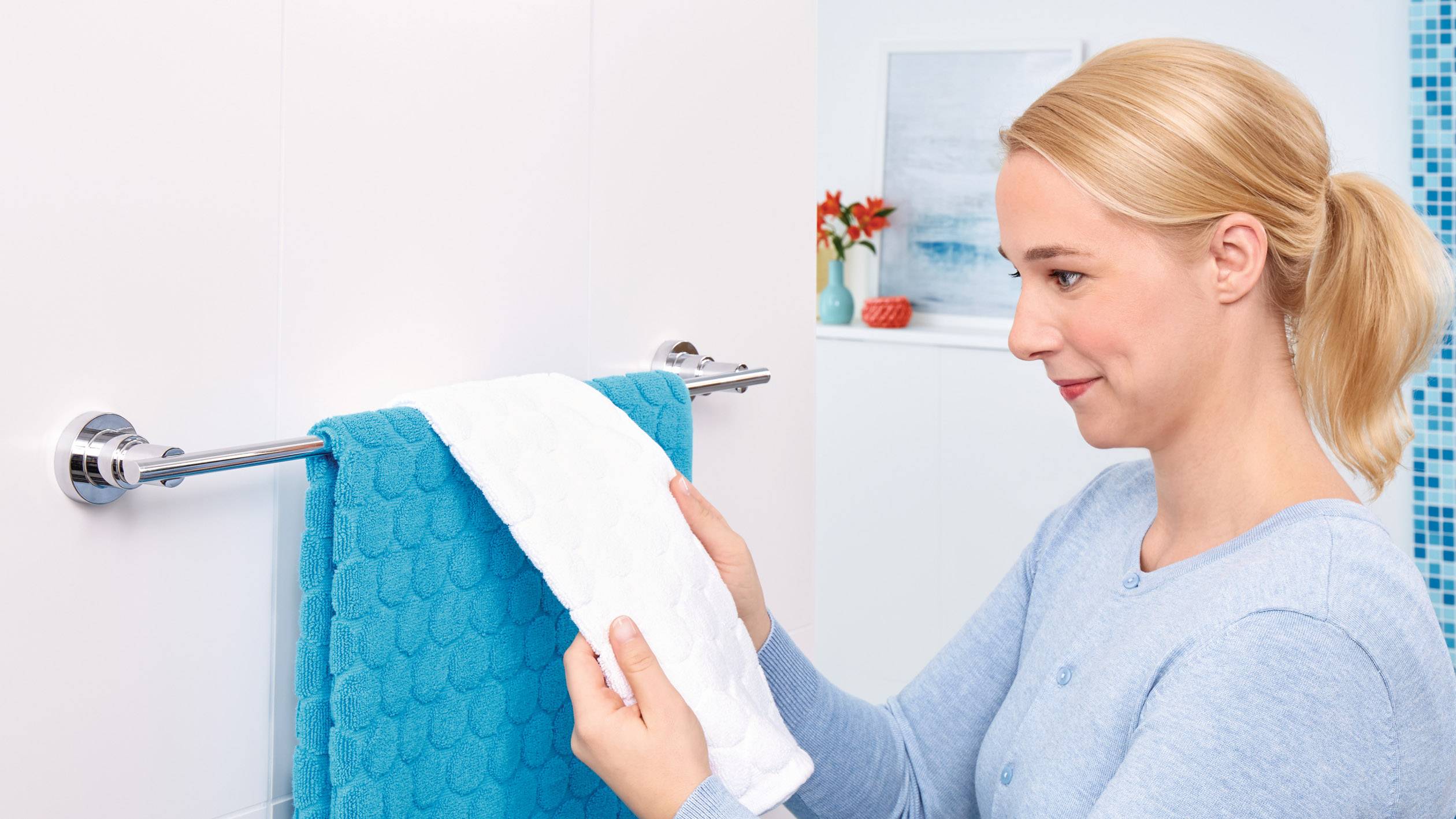 Eine Frau hängt ein weißes Handtuch über ein blaues auf einem Handtuchhalter im Badezimmer auf. Im Hintergrund ist Deko sichtbar.