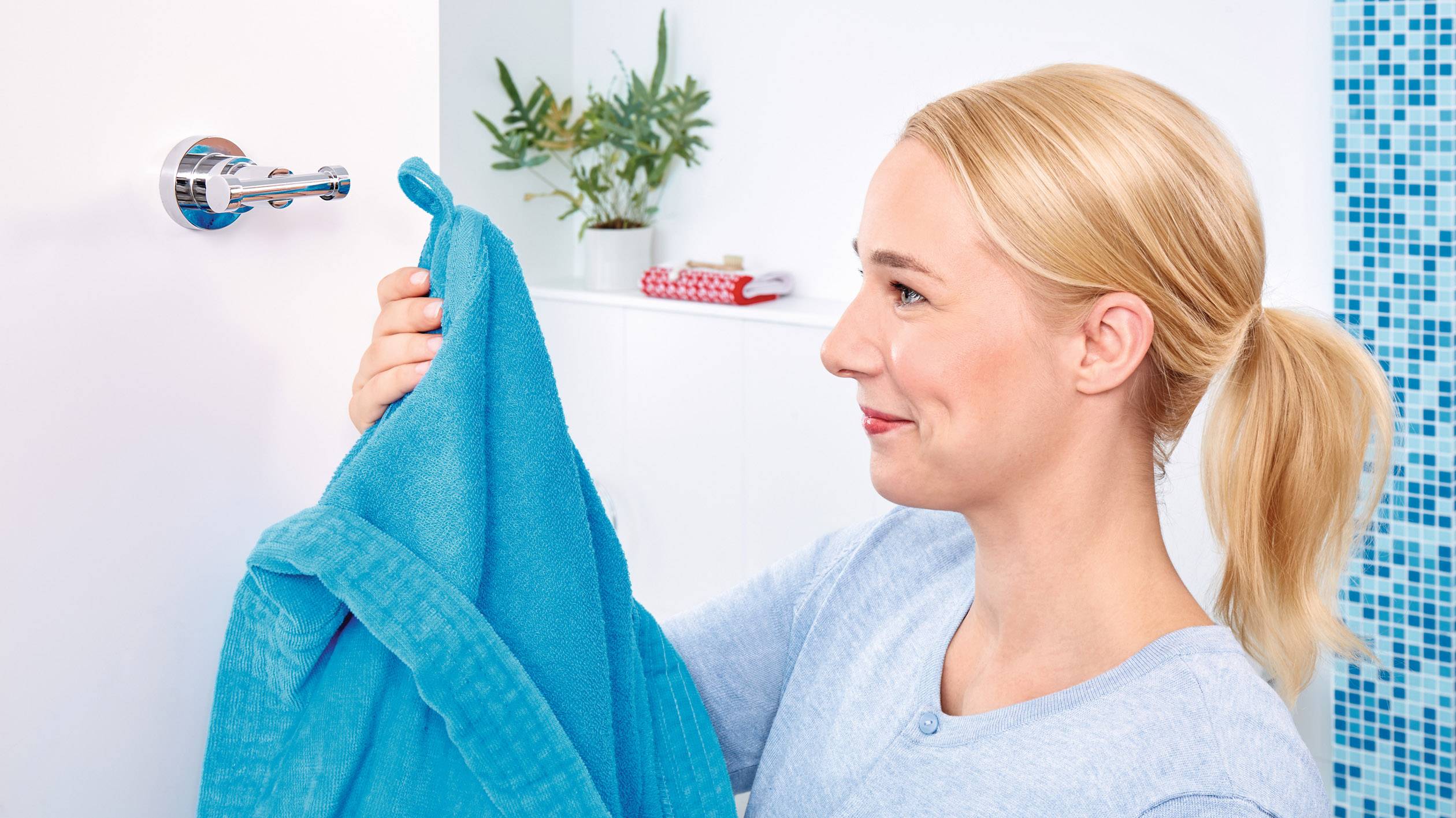 Eine lächelnde Frau hängt ein blaues Handtuch im Badezimmer auf. Im Hintergrund sind Pflanzen und farbige Handtücher auf einem Regal.
