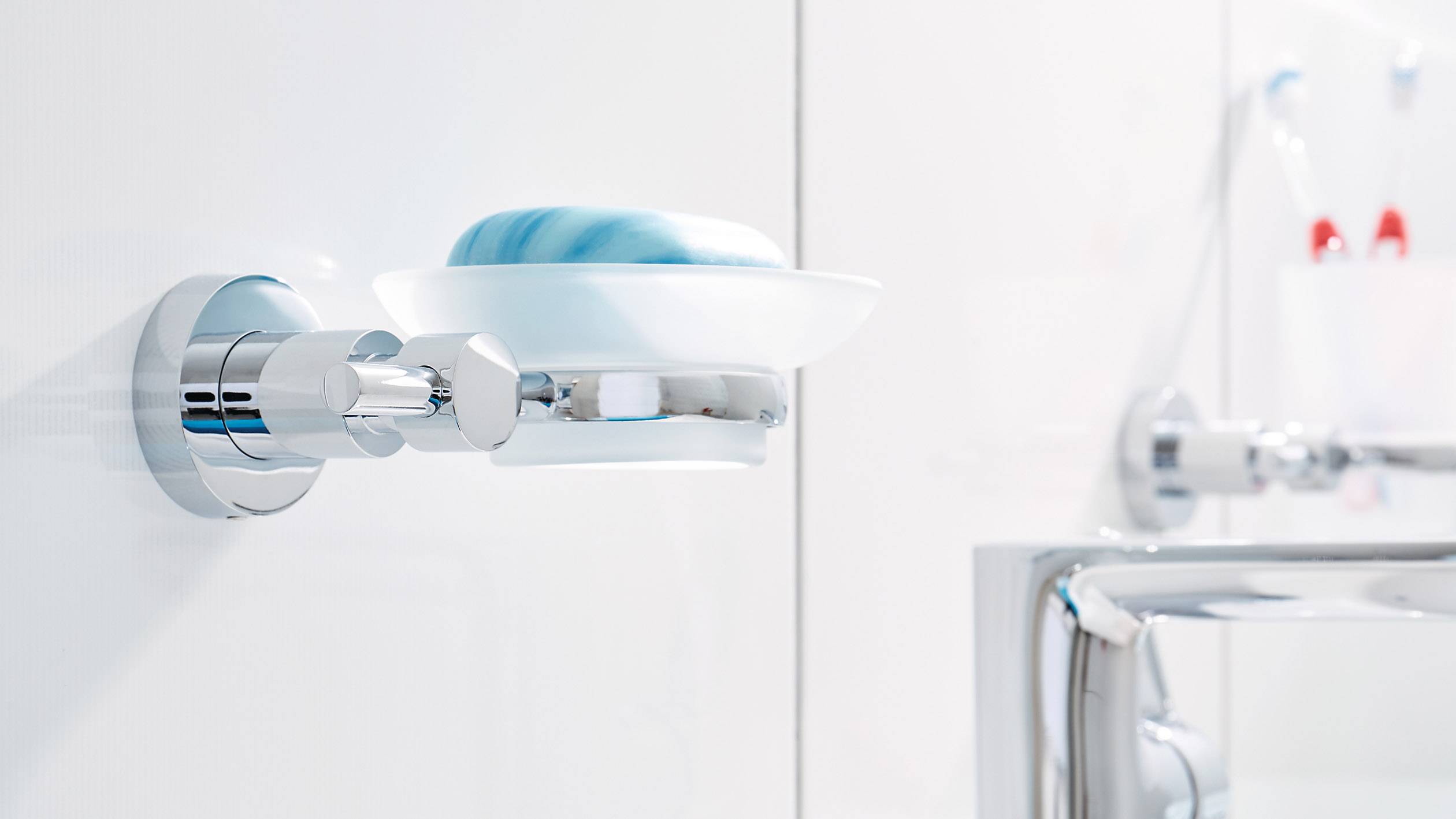 Wandhalterung aus Chrom mit einer weißen Seifenschale, auf der sich ein blau-weißes Stück Seife befindet. Im Hintergrund sind unscharfe Badezimmerzubehörteile zu sehen.