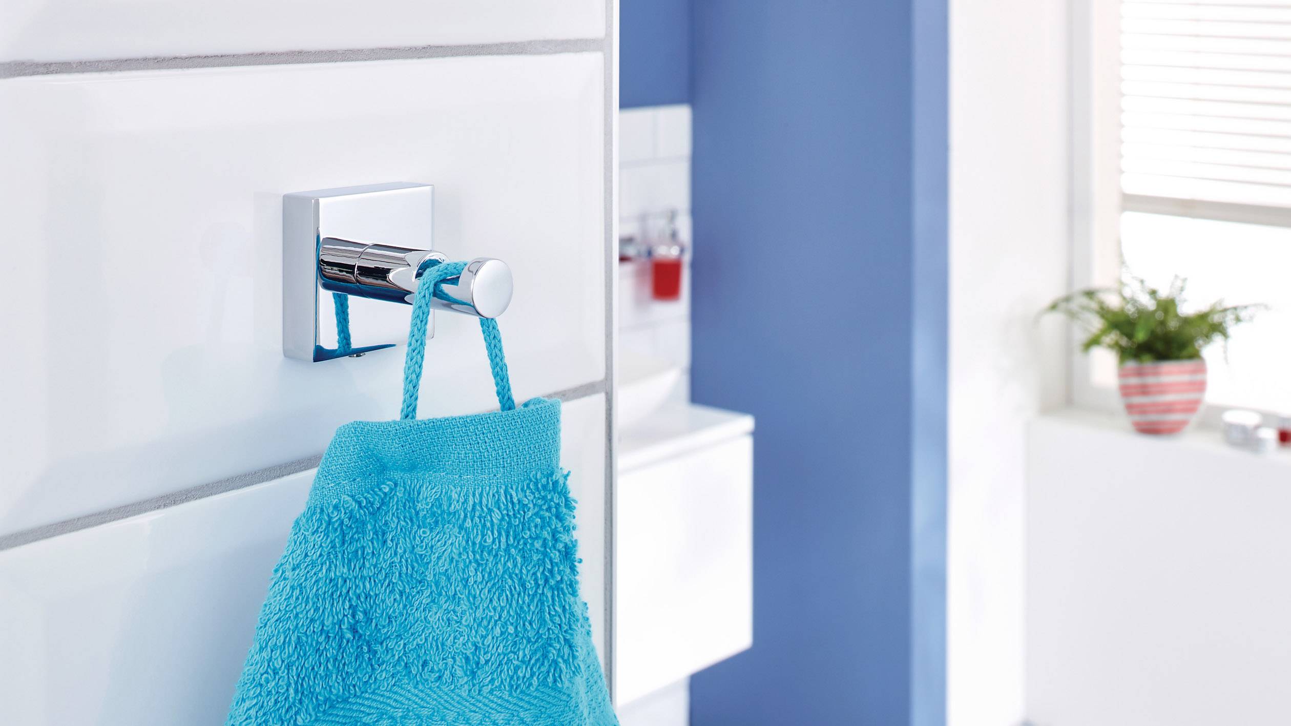 Ein blaues Handtuch hängt an einem metallischen Haken an einer weißen Wand, im Hintergrund ein blau-weißes Badezimmer mit einer Pflanze.