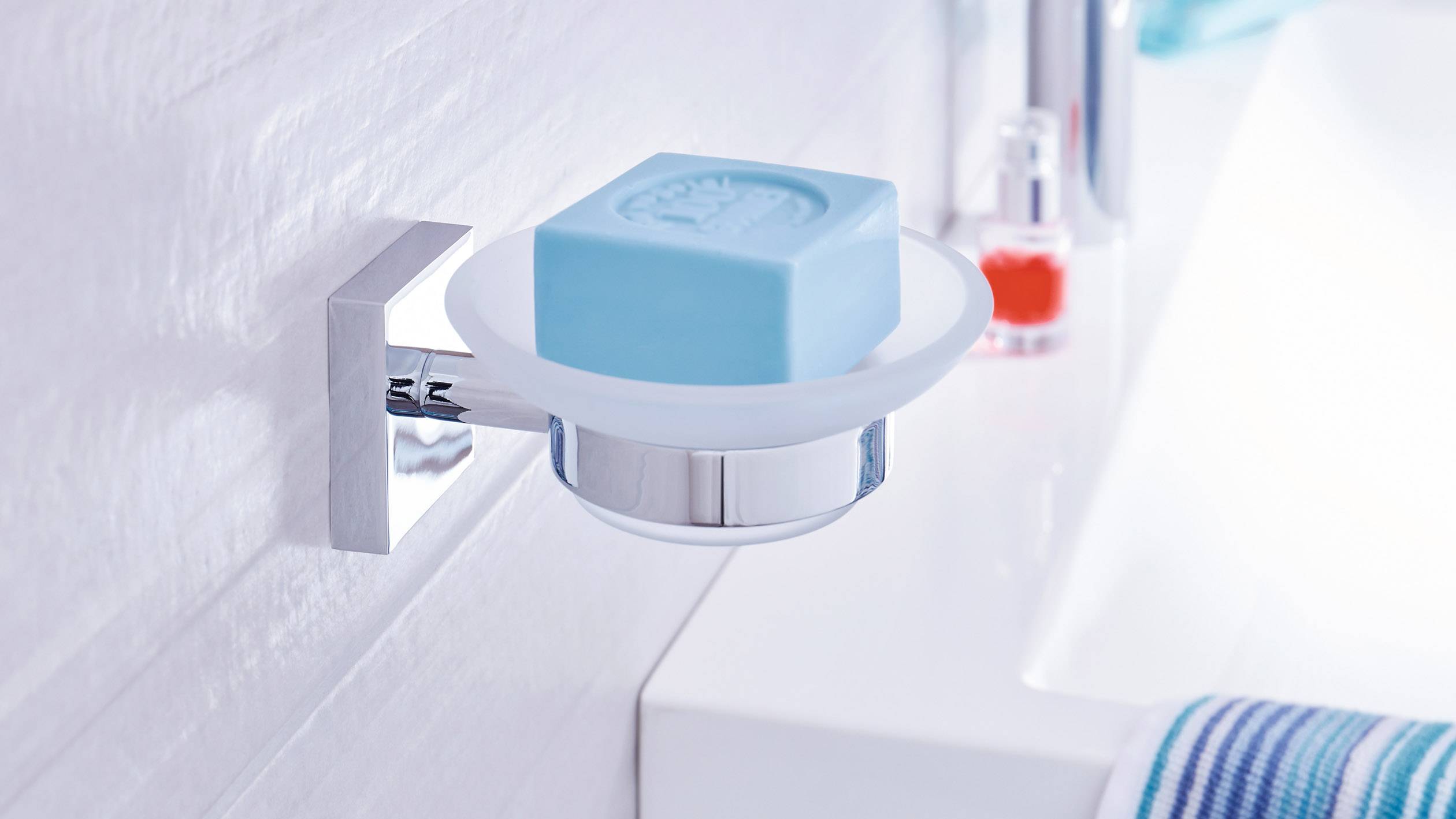 Blaue Seife in einem Halter an einer Badezimmerwand, daneben ein Waschbecken mit Handtuch und ein roter Spender im Hintergrund.