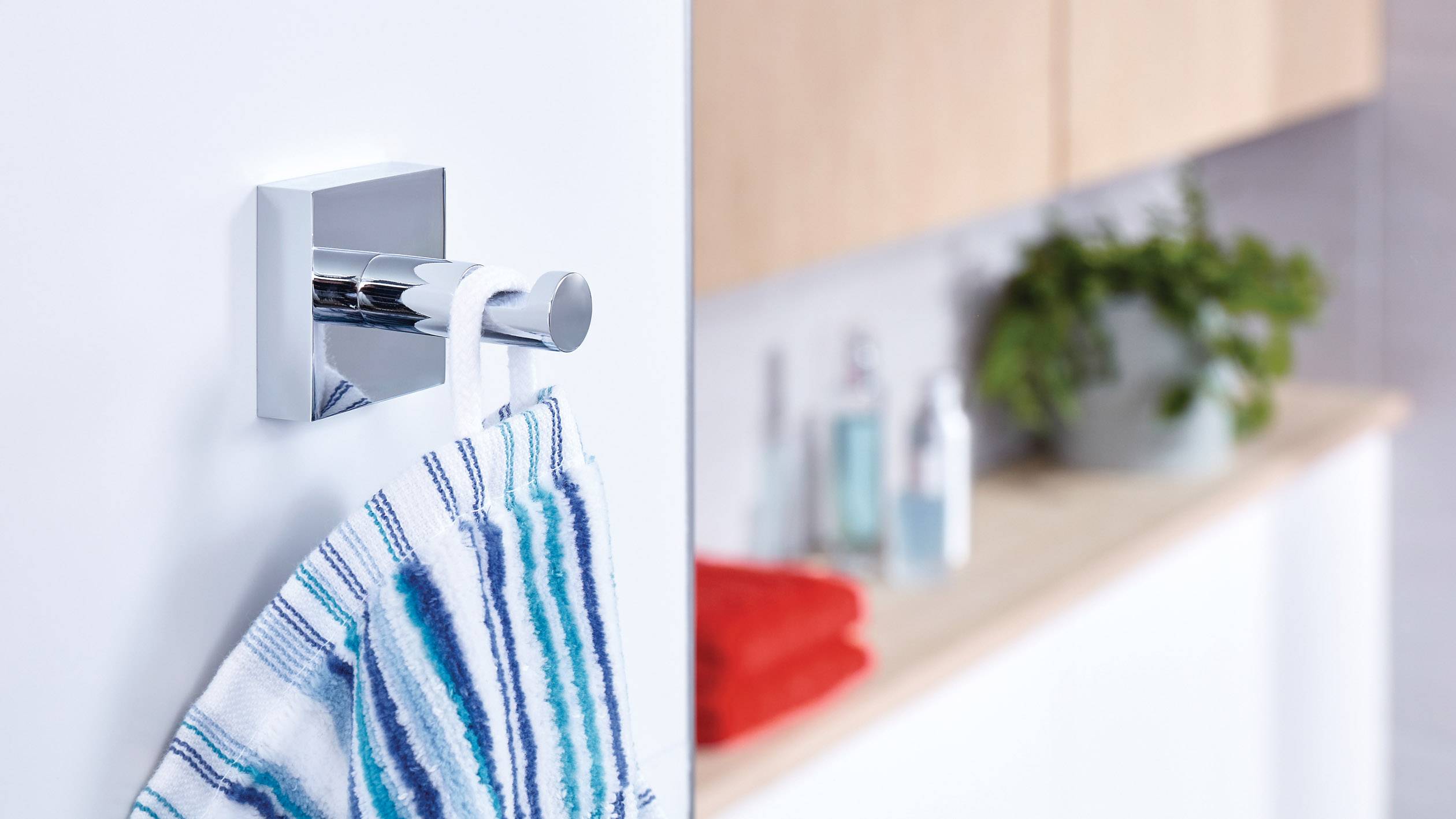 Handtuch hängt an einem modernen silbernen Wandhaken im Badezimmer. Im Hintergrund sind Pflegeprodukte und ein rotes Handtuch.