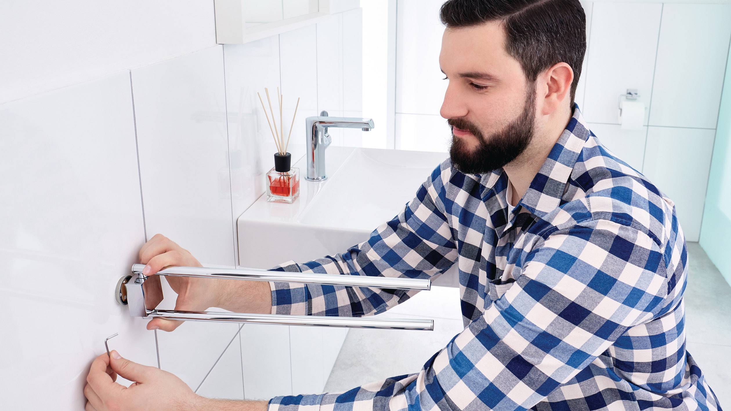 Ein Mann in einem karierten Hemd repariert im Badezimmer eine Wasserleitung mit Werkzeug. Im Hintergrund ist ein Waschbecken zu sehen.