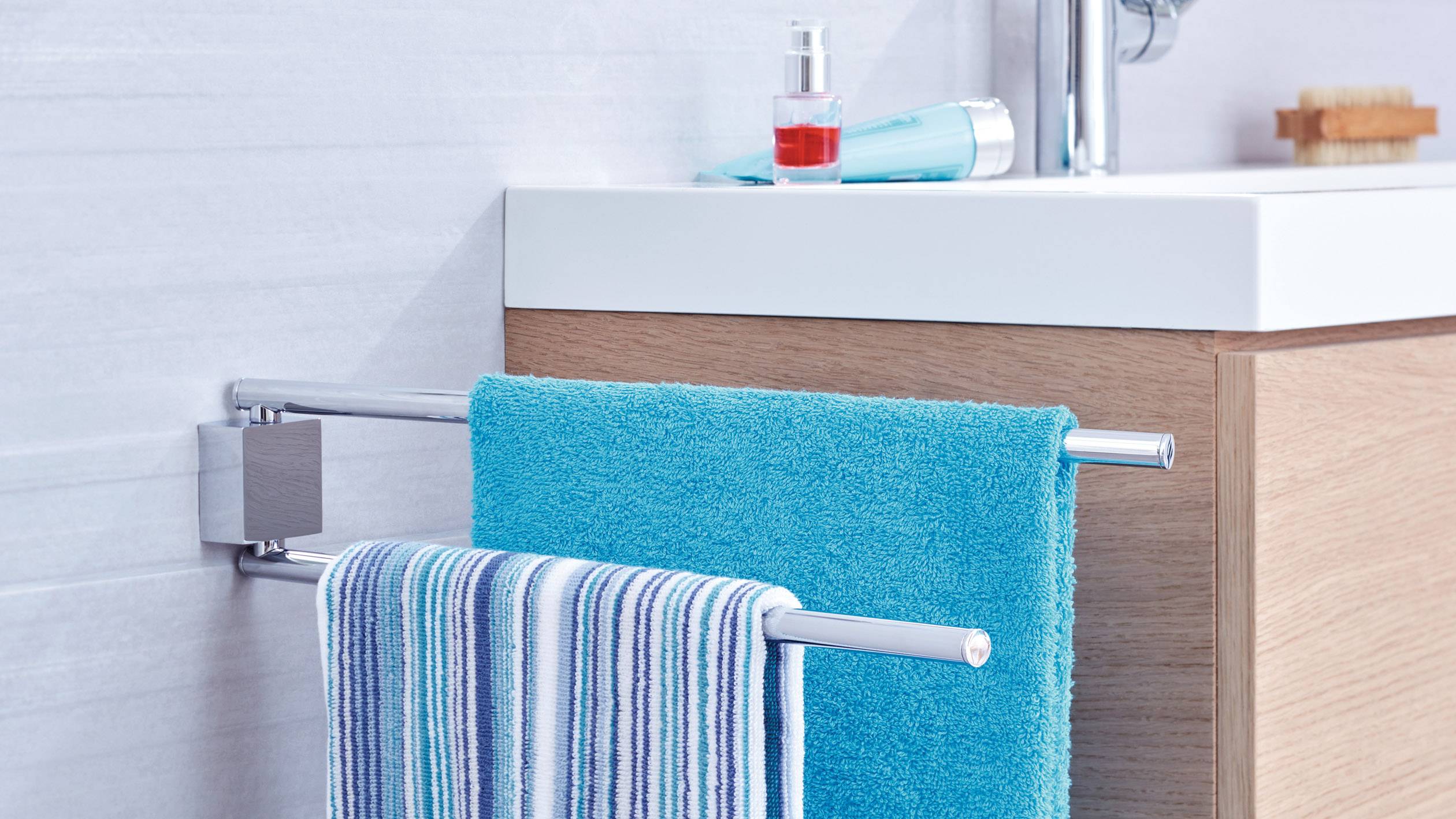 Ein Badezimmer mit einem Handtuchhalter, darauf ein gestreiftes und ein türkisfarbenes Handtuch. Im Hintergrund Waschbecken mit Seifenspender.