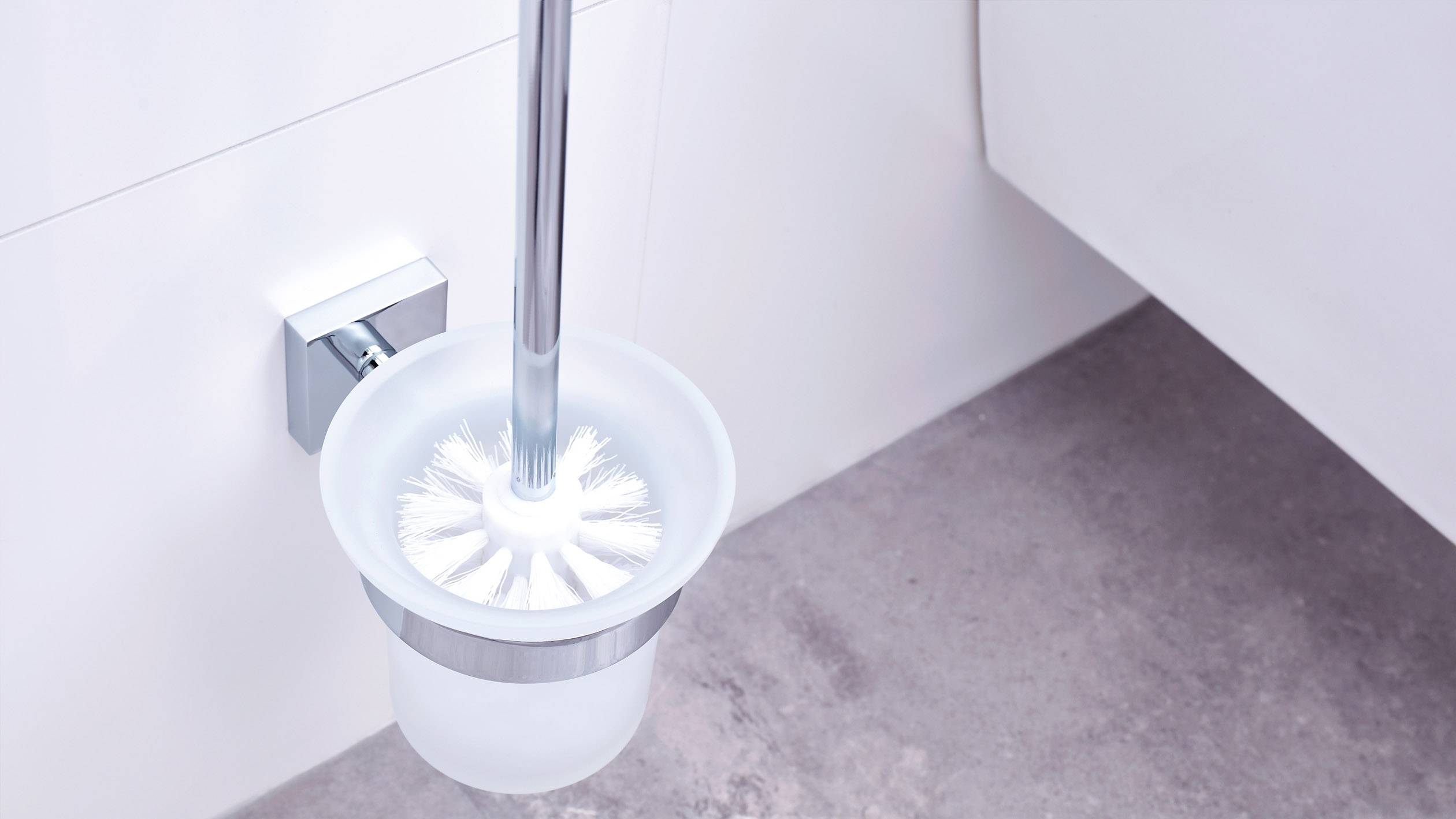 Eine Toilettenbürste in einem weißen Behälter ist an der Wand eines Badezimmers angebracht. Der Boden ist grau gefliest.