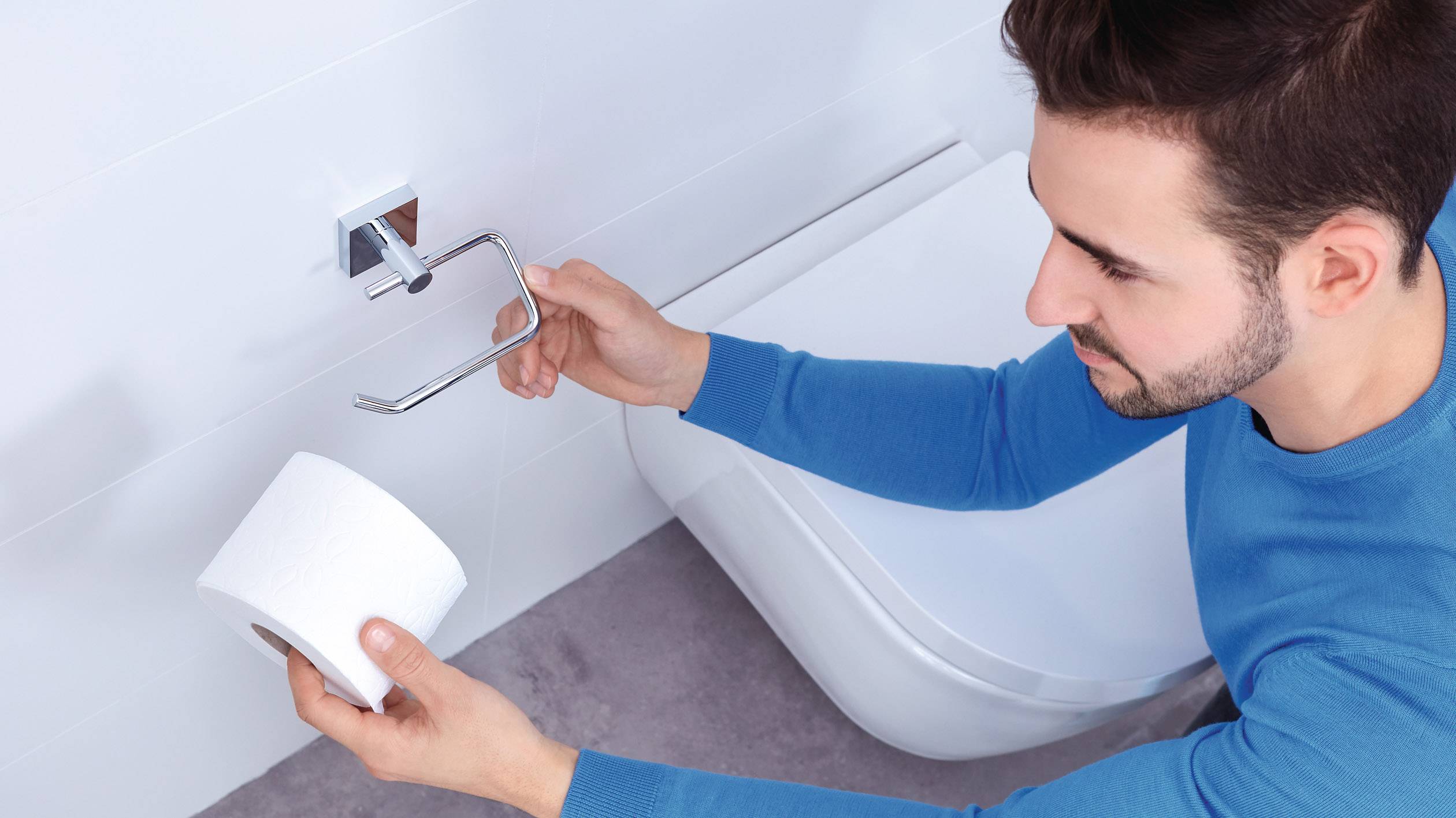 Ein Mann in einem blauen Pullover montiert einen Toilettenpapierhalter an einer Badezimmerwand.