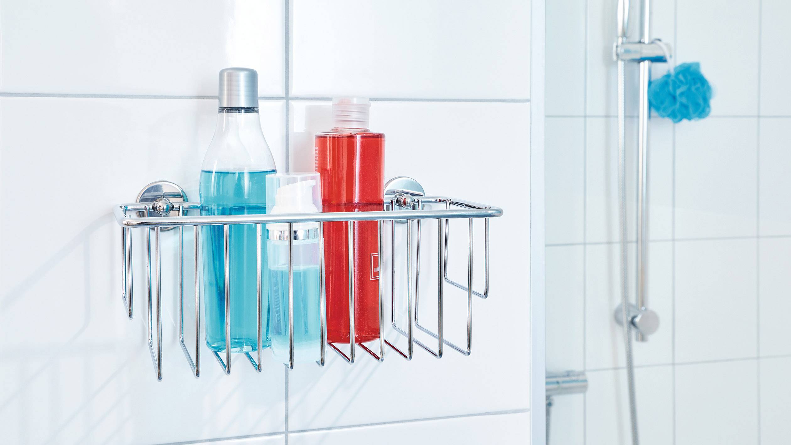 Duschkorb aus Metall mit zwei bunten Flaschen, montiert an einer gefliesten Badezimmerwand neben einer Dusche.