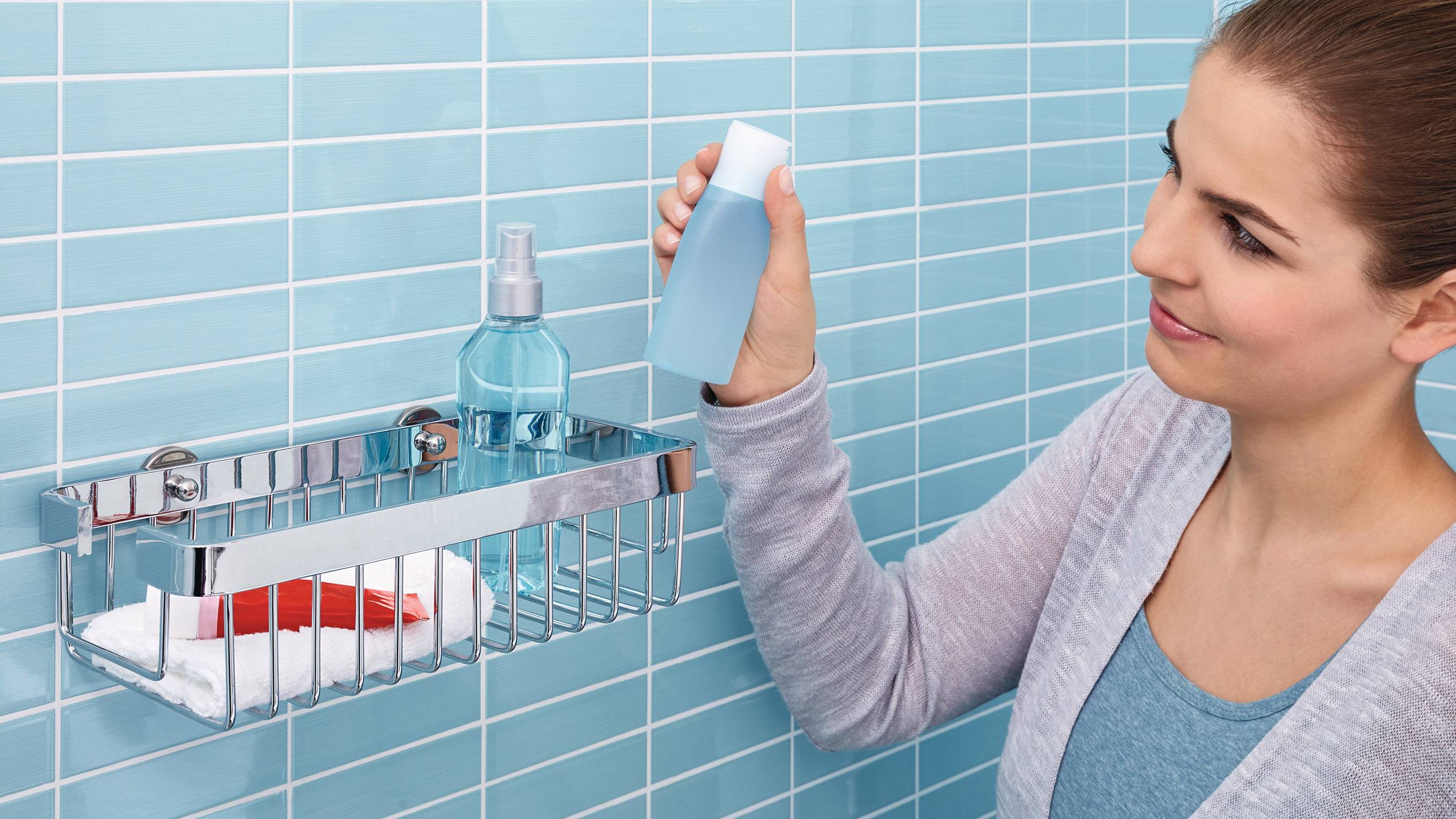 Eine Person stellt eine blaue Flasche in ein Metallregal im Badezimmer. Im Regal befinden sich bereits ein roter Kamm und eine Sprühflasche.