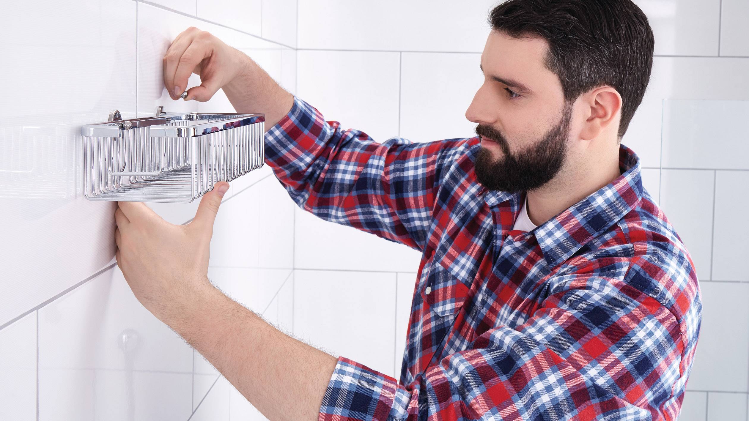 Ein Mann montiert ein Metallregal an eine geflieste Badezimmerwand. Er trägt ein kariertes Hemd und benutzt ein Werkzeug.