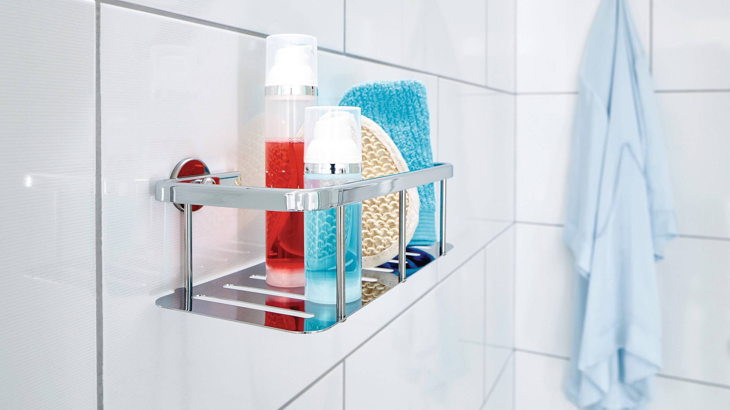Ein Badezimmerregal mit Duschgel, Schwamm und weiteren Pflegeprodukten an einer weißen gefliesten Wand. Ein blaues Handtuch hängt daneben.