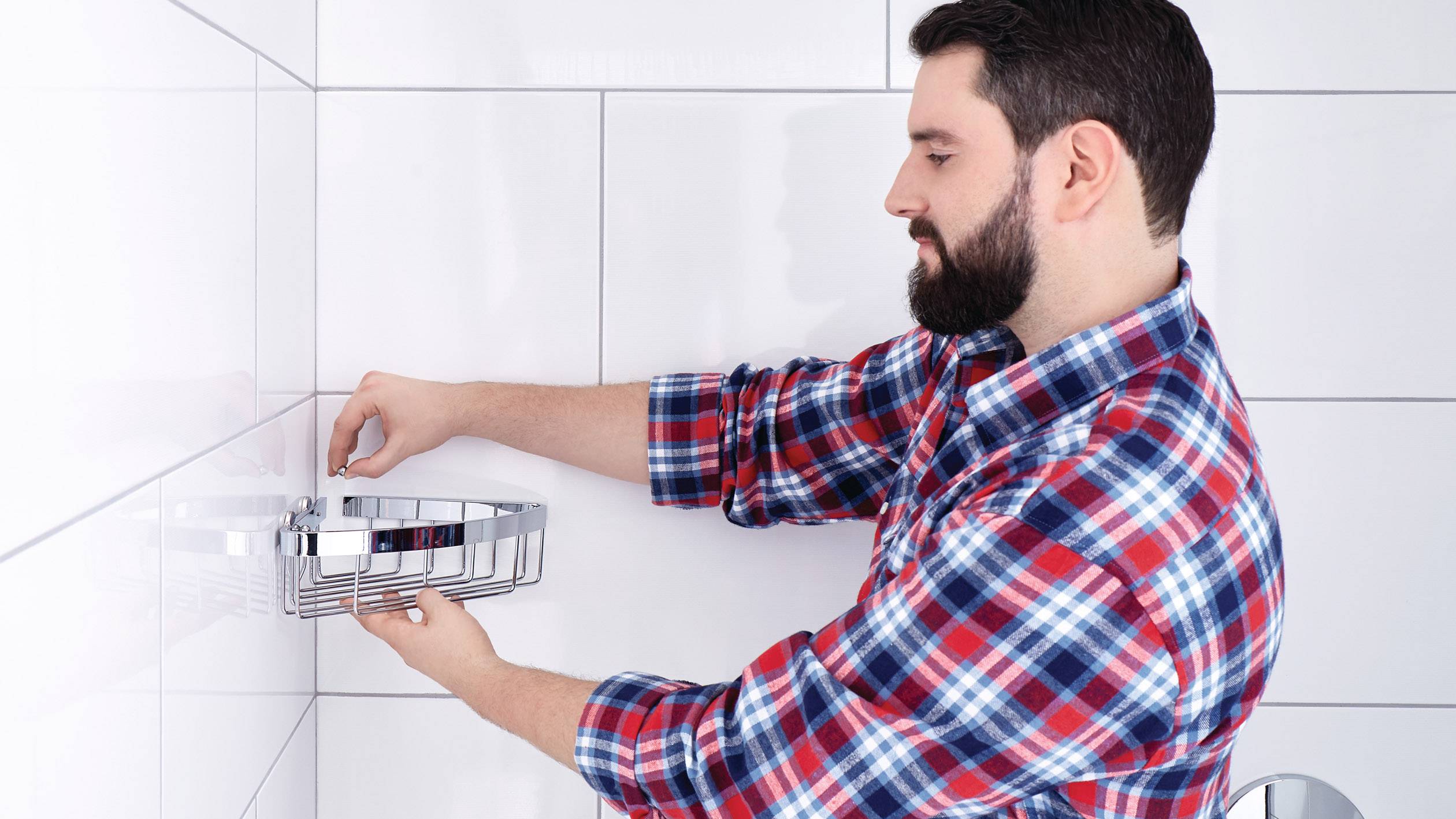 Ein Mann in kariertem Hemd montiert ein Duschregal an einer weißen Fliesenwand.