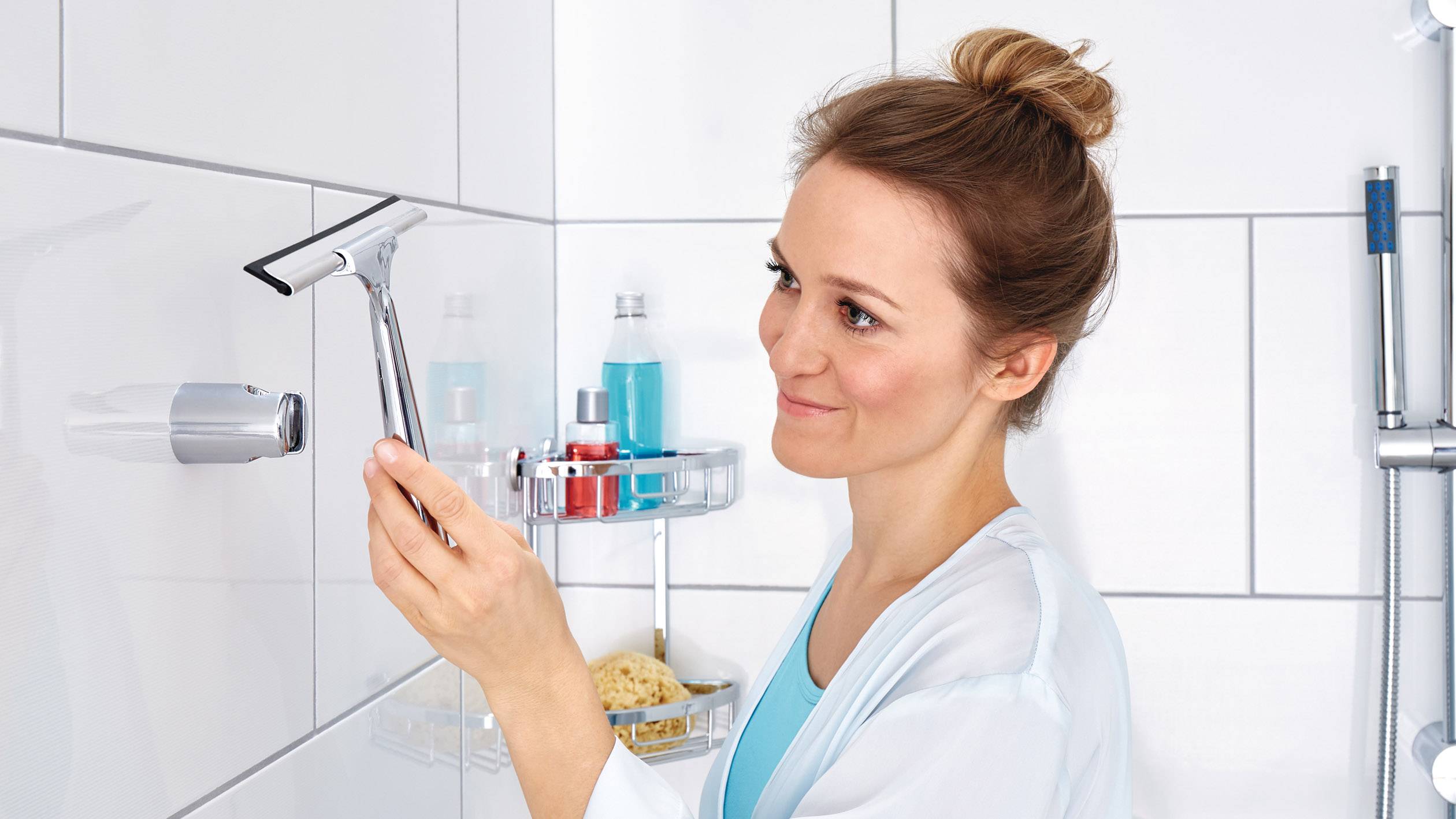 Eine Frau reinigt Fliesen im Badezimmer mit einem Abzieher. Neben ihr befinden sich Hygieneprodukte auf einem Duschregal.