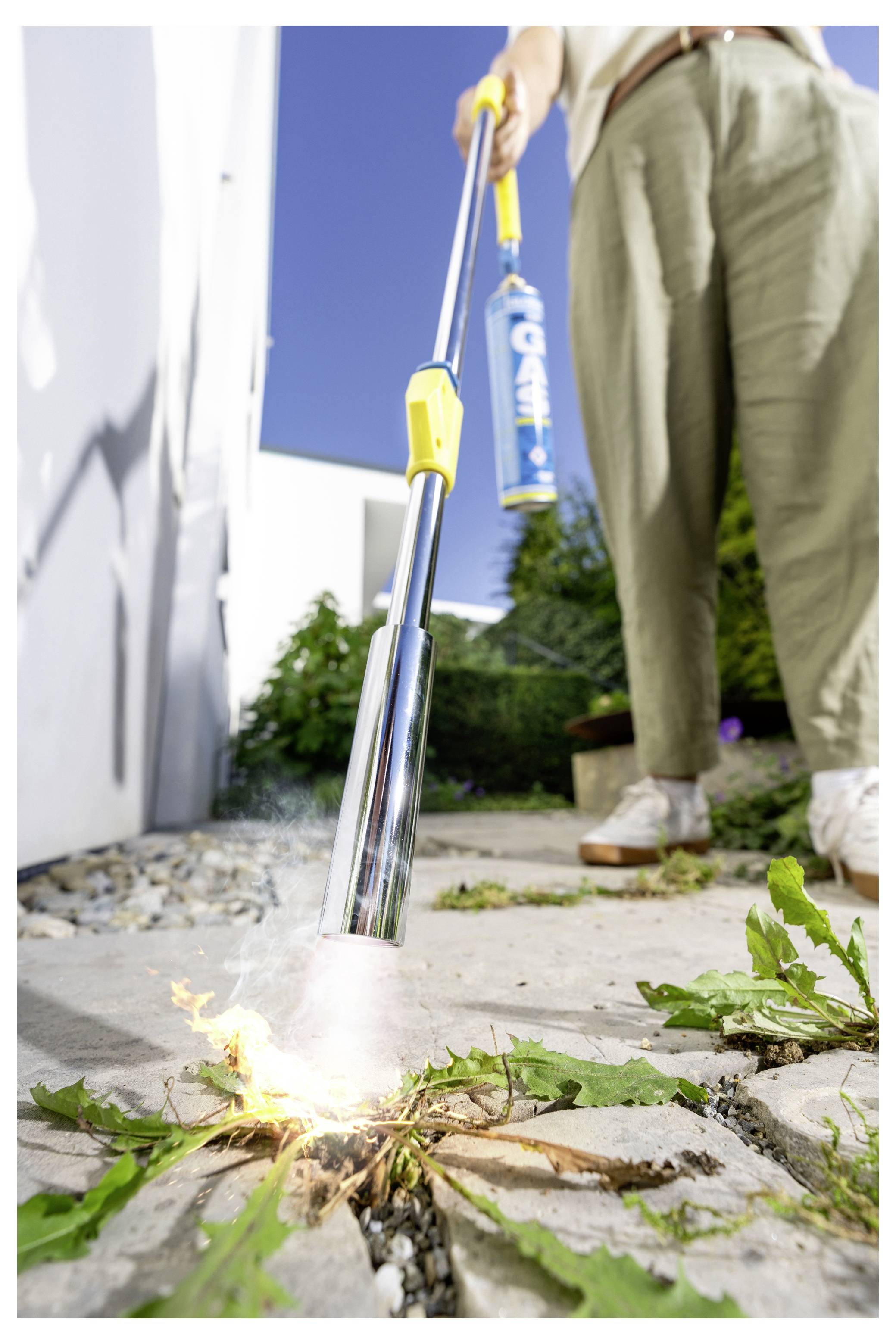 Person, die eine tragbare Fackel benutzt, um Unkraut in Rissen zwischen Gehwegplatten neben einem Gebäude zu verbrennen.