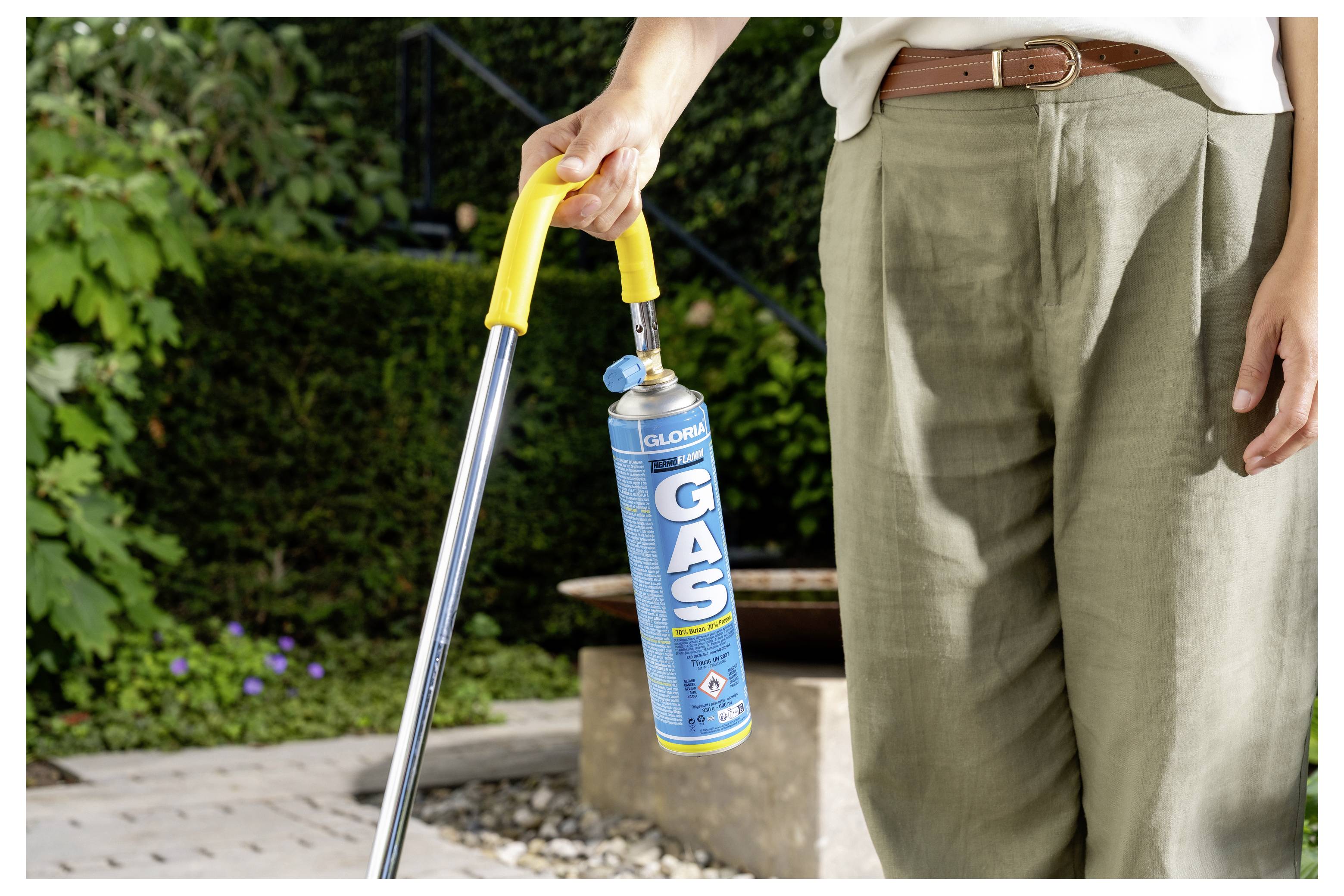 Eine Person hält eine Gaskartusche, die an einem tragbaren Brenner befestigt ist, und steht im Freien in einem Garten- oder Terrassenbereich.