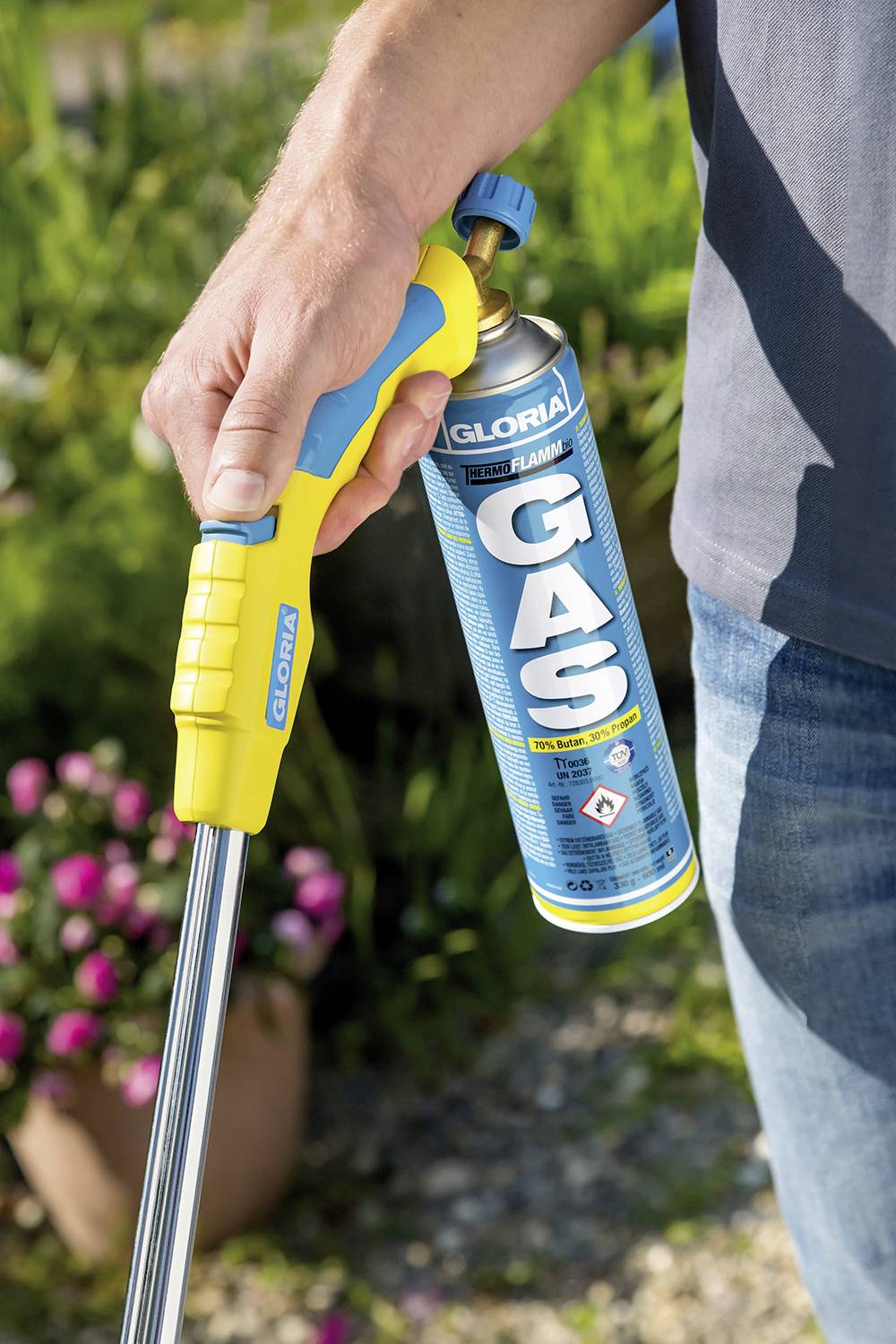 Eine Person hält ein gelbes und blaues Gasgerät mit einer Gaskartusche von 'GLORIA'. Im Hintergrund sind Pflanzen und Blumen.