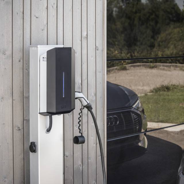 Ladestation mit angeschlossenem Kabel an der Wand montiert, vor einem geparkten Auto im Freien, bei Tageslicht.
