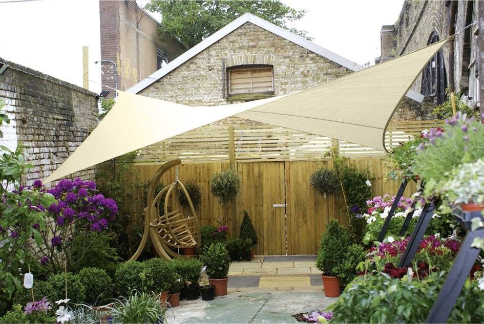 Ein Innenhof mit Holzzaun, umgeben von Topfpflanzen und Blumen. Zwei Sonnensegel spenden Schatten. Links steht ein Hängekorb aus Holz.