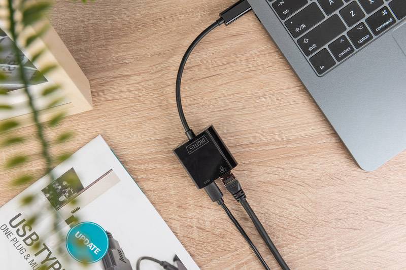 Drei-in-eins-USB-C-Hub verbindet Laptop mit mehreren Geräten auf Holztisch. Neben dem Hub liegt eine Zeitschrift und eine Pflanze.