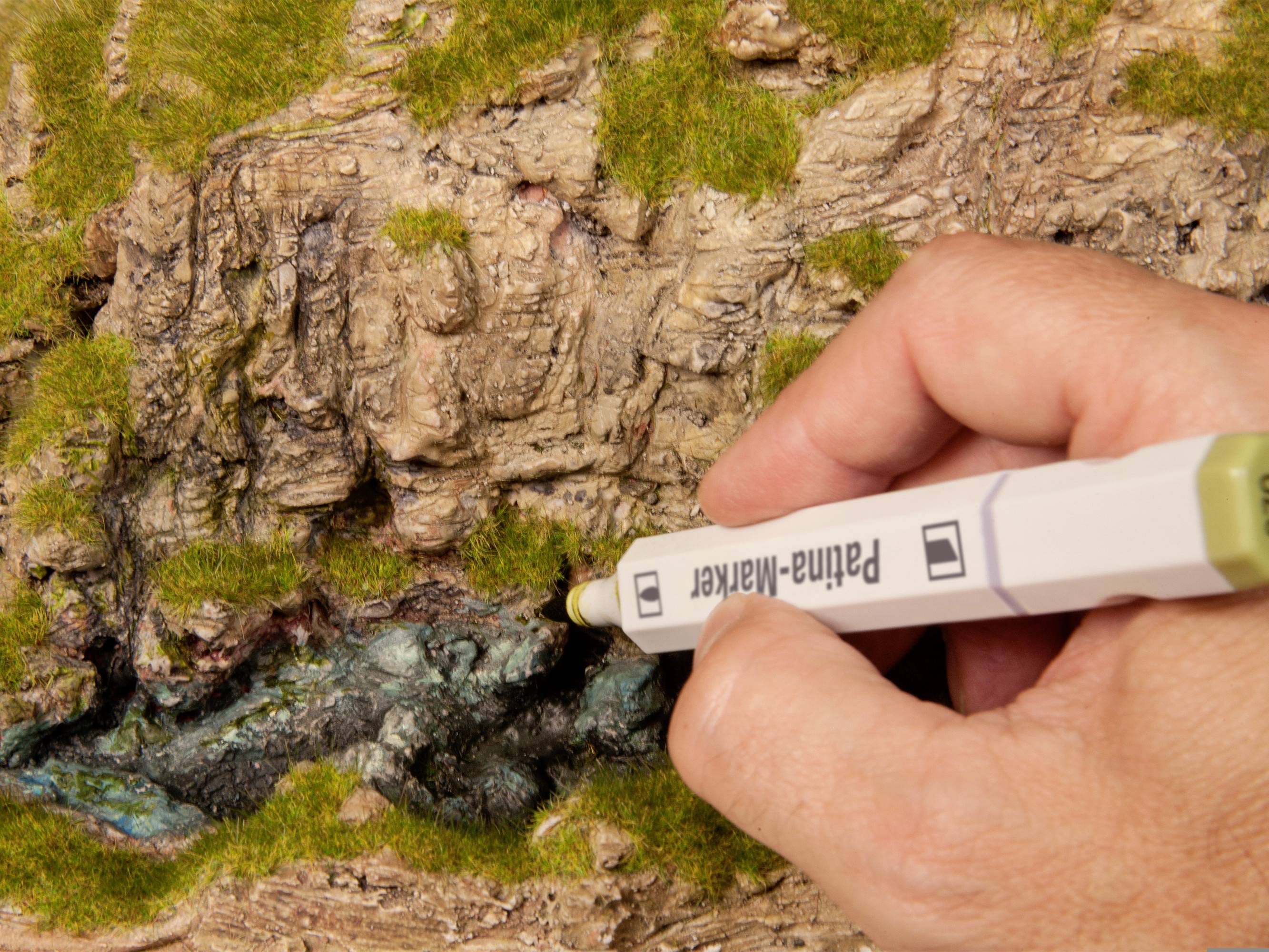 Eine Hand hält einen Marker und bemalt ein detailliertes Modell eines felsigen Hügels mit Moos und Gras.