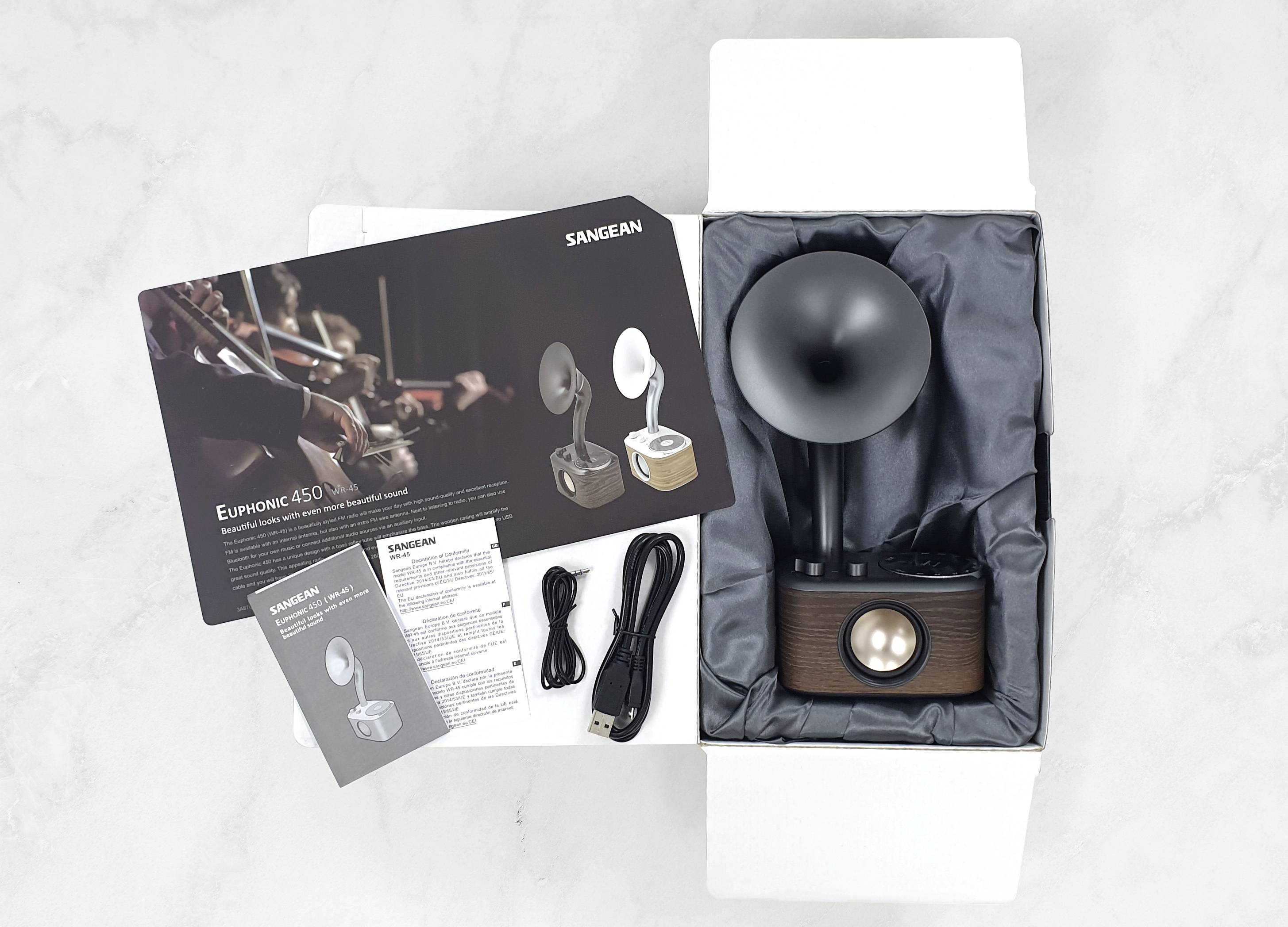Ein offener Karton mit einem Retro-Lautsprecher, USB-Kabel, Audiokabel und Broschüren. Markentext 'SANGEAN' sichtbar.