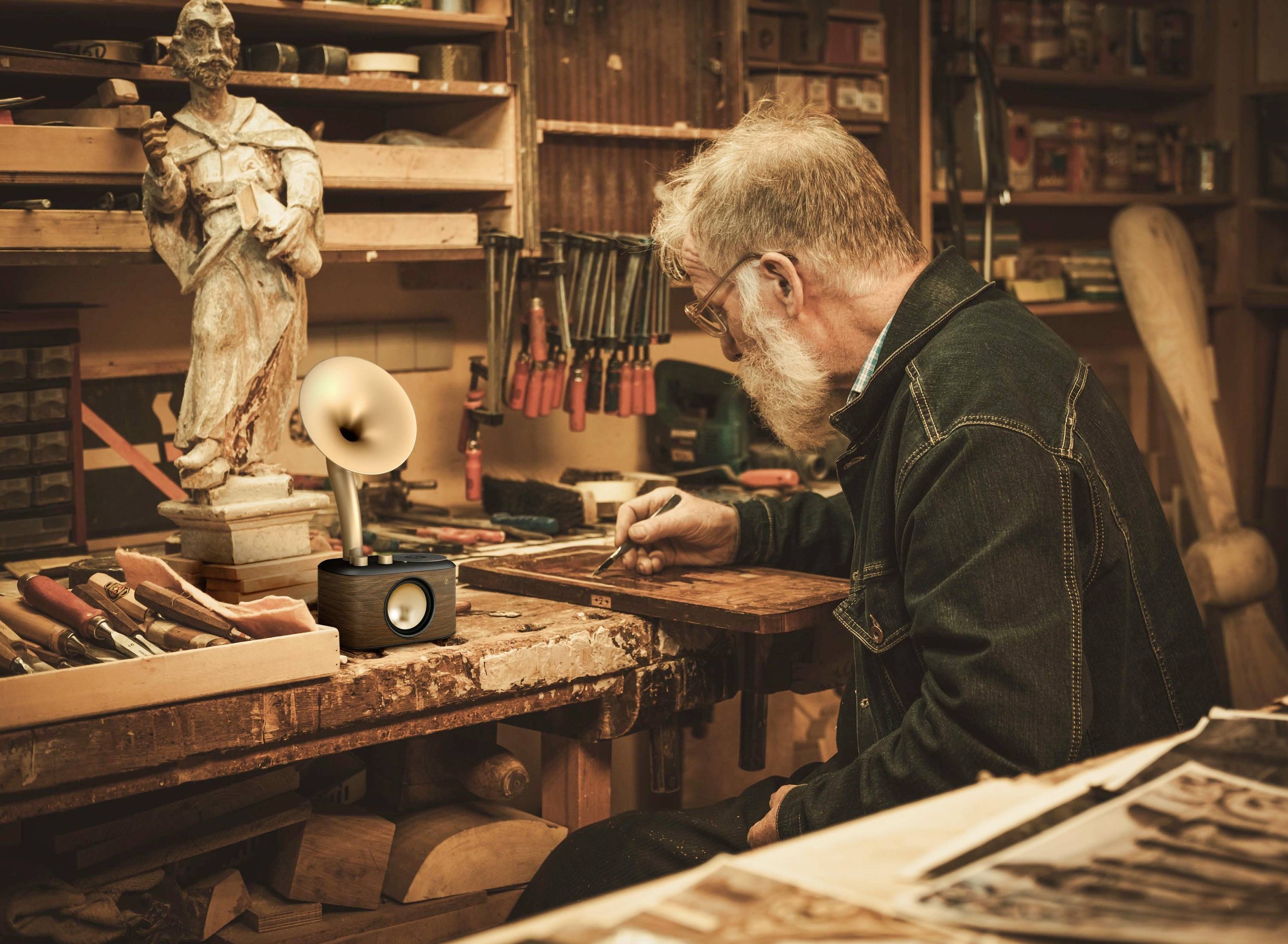 Ein Mann mit grauem Haar und Bart arbeitet an einem Tisch in einer Werkstatt, umgeben von Werkzeugen und einer Skulptur.