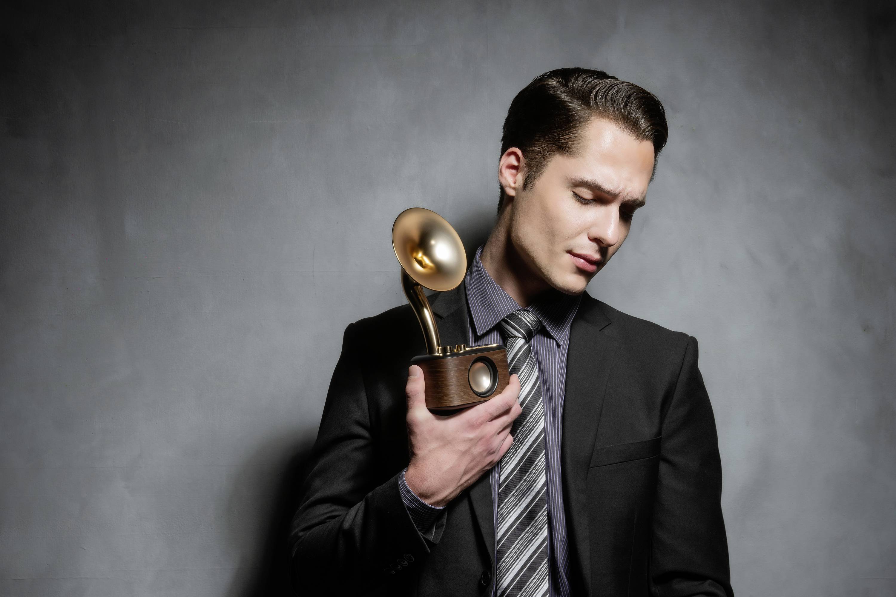 Ein Mann im Anzug hält eine Grammophon-Auszeichnung. Er schaut nach unten und steht vor einem neutralen Hintergrund.