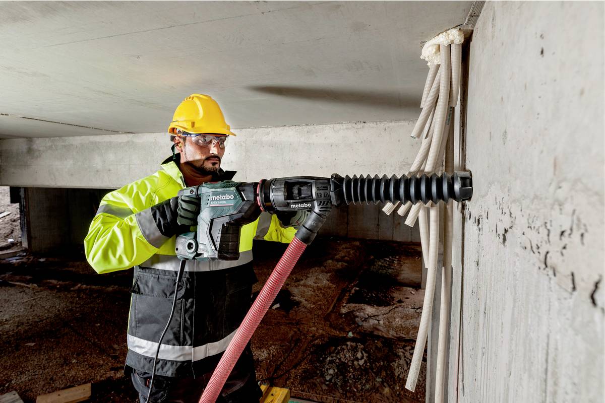 Ein Bauarbeiter in Schutzkleidung bohrt mit einer Bohrmaschine ein Loch in eine Betonwand. Im Hintergrund sind Baustellenstrukturen sichtbar.