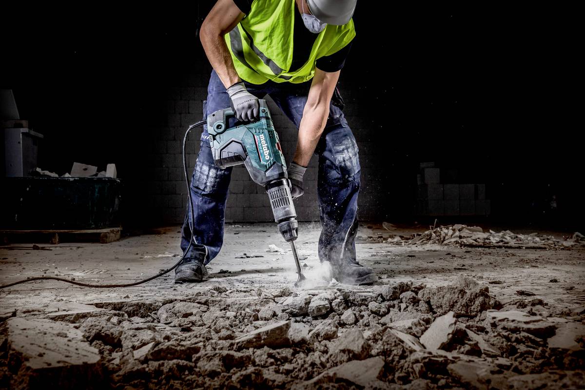 Ein Bauarbeiter mit Schutzweste und Maske benutzt einen Presslufthammer, um Beton auf einer Baustelle aufzubrechen.