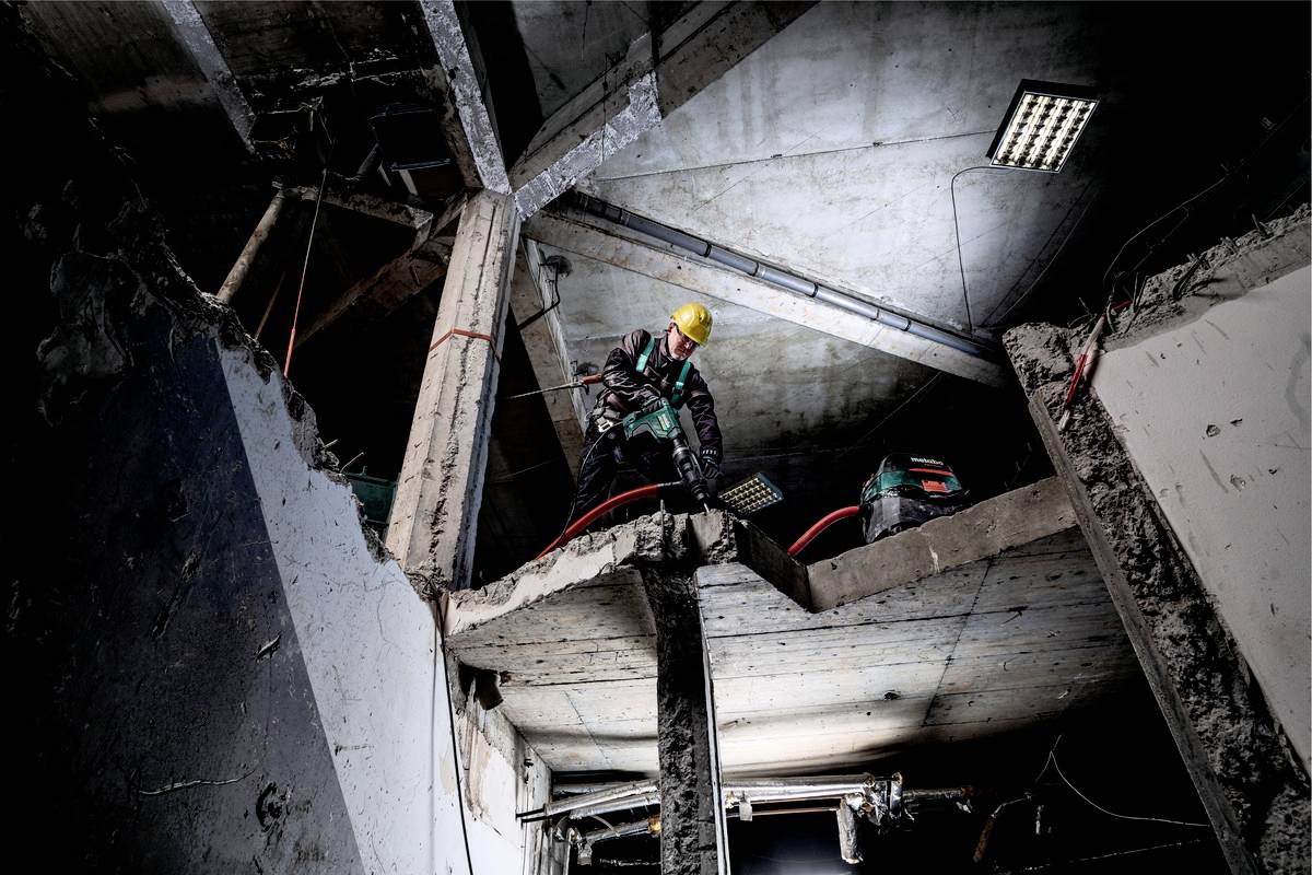 Arbeiter mit Schutzhelm steht auf einer Baustelle mit Betontrümmern und verwendet ein Werkzeug, um Beton zu bohren oder zu schneiden.