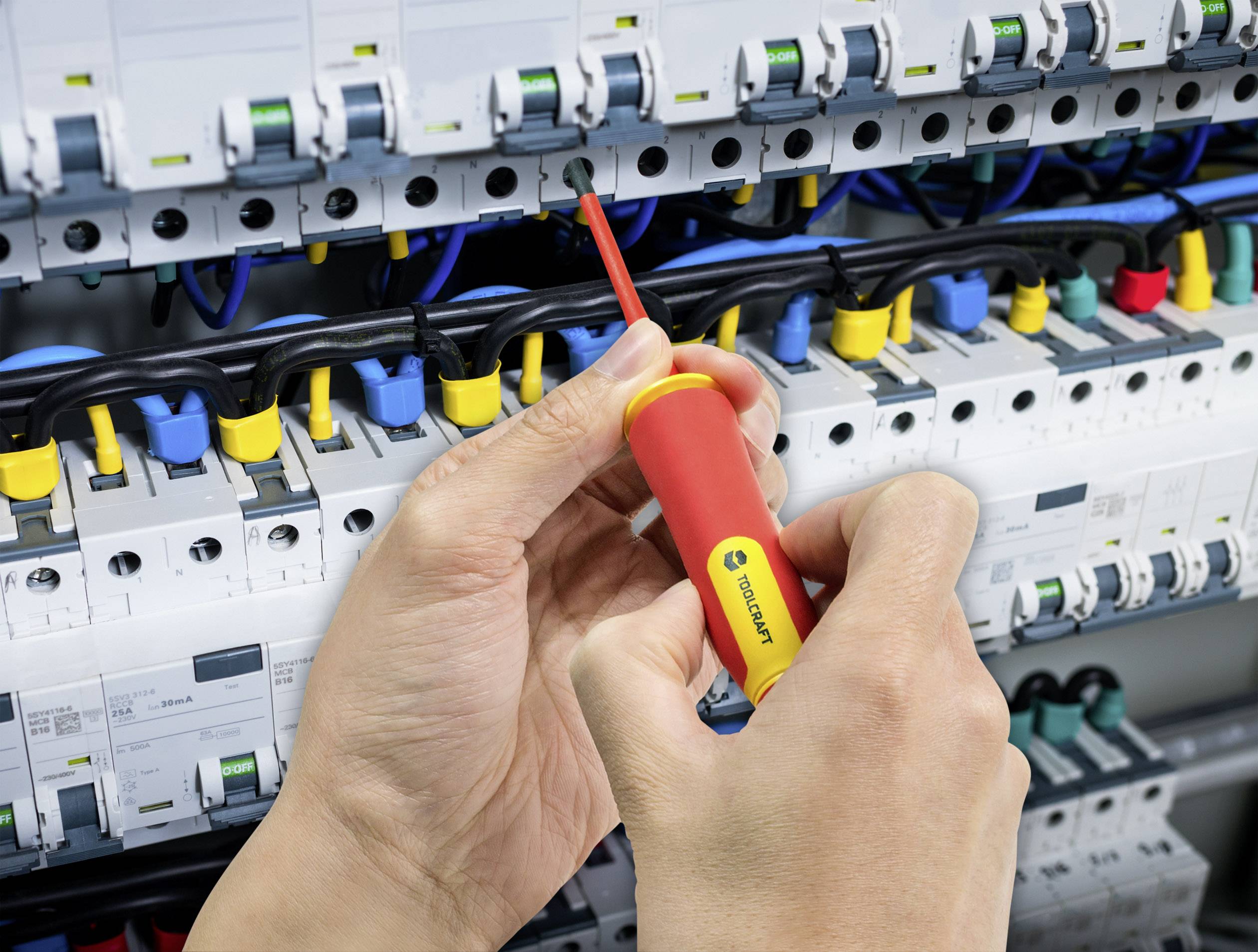 Eine Person benutzt einen Schraubendreher, um an einer elektrischen Schalttafel Kabel anzuschließen. Verschiedene farbige Kabel sind sichtbar.