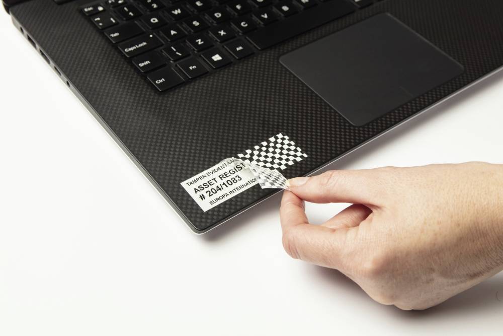 Eine Hand entfernt ein Sicherheitsaufkleber von einem Laptop. Der Aufkleber zeigt eine Seriennummer und ein Rautenmuster, das beim Ablösen sichtbar wird.