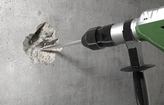 Ein Bohrer durchbricht eine Betonwand. Der Bohrer erzeugt Staub und Bruchstücke. Die Szene zeigt eine handwerkliche Tätigkeit.