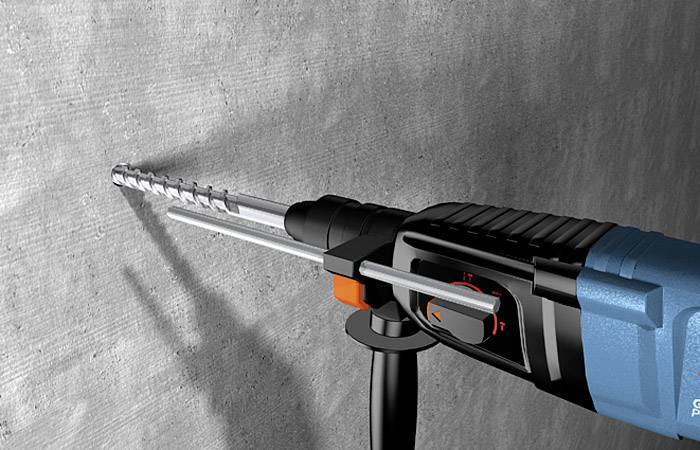 Ein Bohrhammer bohrt ein Loch in eine Betonwand. Die Maschine ist blau-schwarz mit einem Metallbohrer und einem Griff an der Seite.