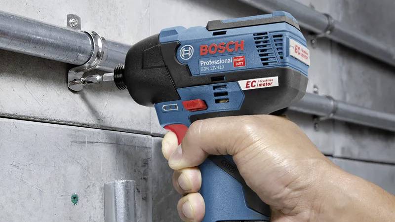 Eine Hand, die einen professionellen Bosch-Elektroschrauber verwendet, um Schrauben an einem Metallrohr zu befestigen, das an einer Betonwand angebracht ist.