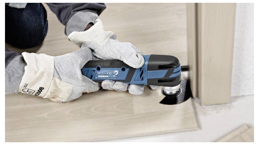 Eine Person mit Handschuhen verwendet ein blaues, tragbares oszillierendes Werkzeug, um Holzbodenbelag in der Nähe des Türrahmens zu schneiden.