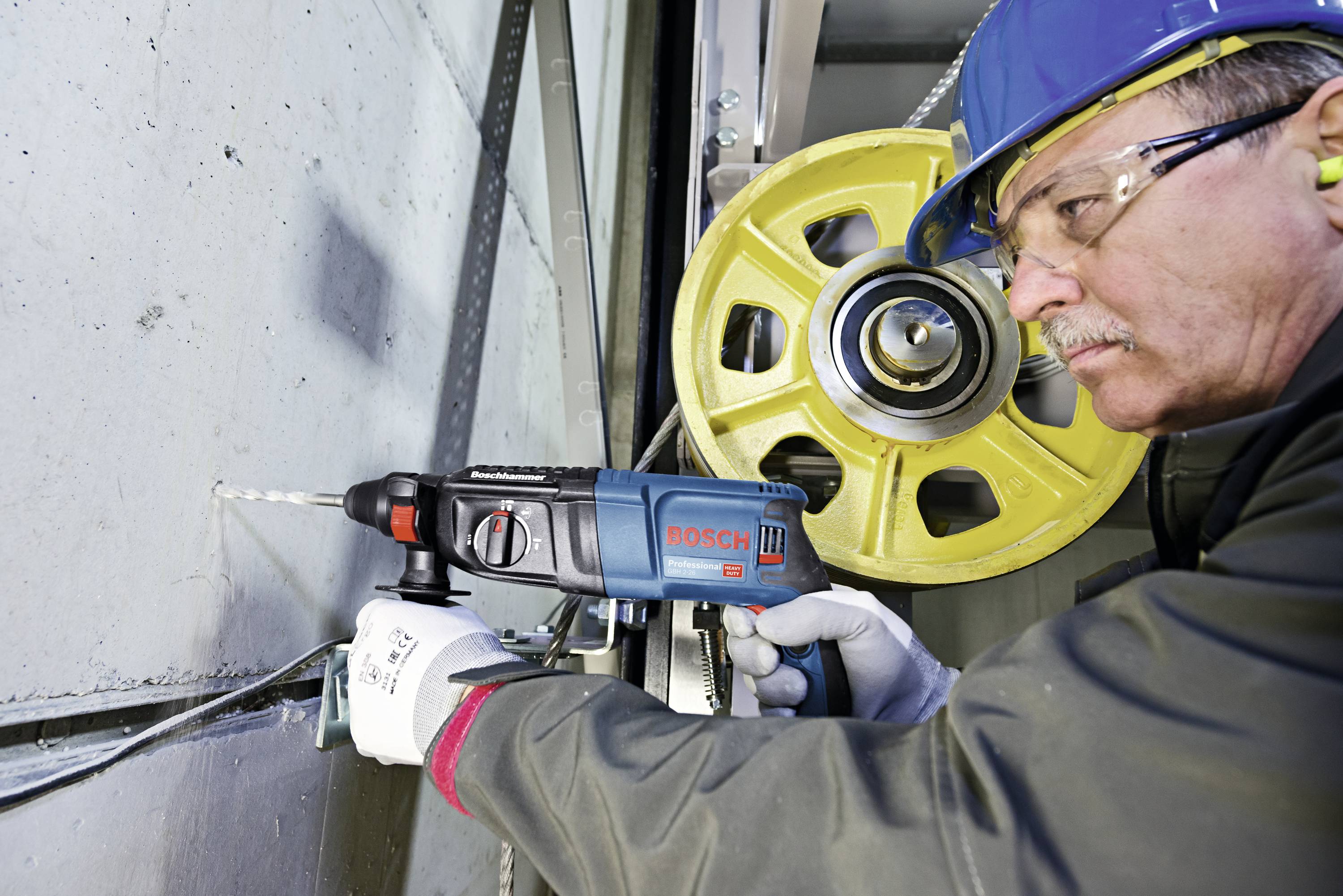 Ein Arbeiter mit Schutzhelm und Handschuhen bohrt mit einer blauen Bosch-Bohrmaschine in eine Wand, industrielle Umgebung im Hintergrund.
