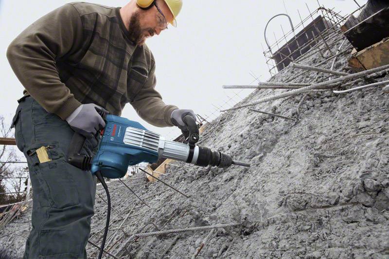 Arbeiter mit Schutzhelm und Ohrenschützern bedient einen Bohrhammer, um Beton auf einer Baustelle zu bearbeiten.