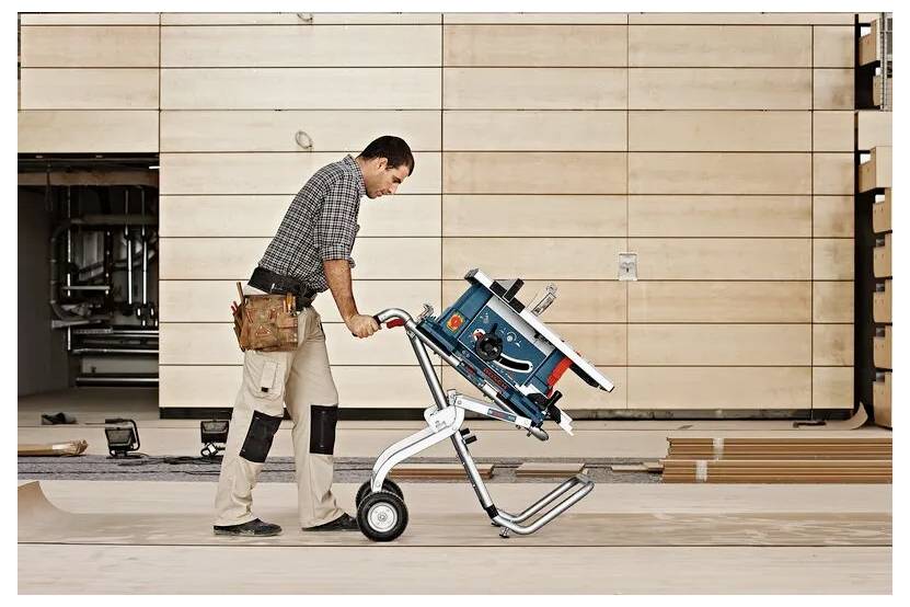 Eine Person schiebt eine tragbare Tischkreissäge auf Rädern über einen Holzboden in einer Werkstattumgebung, wobei sie einen Werkzeuggürtel und Arbeitshosen trägt.