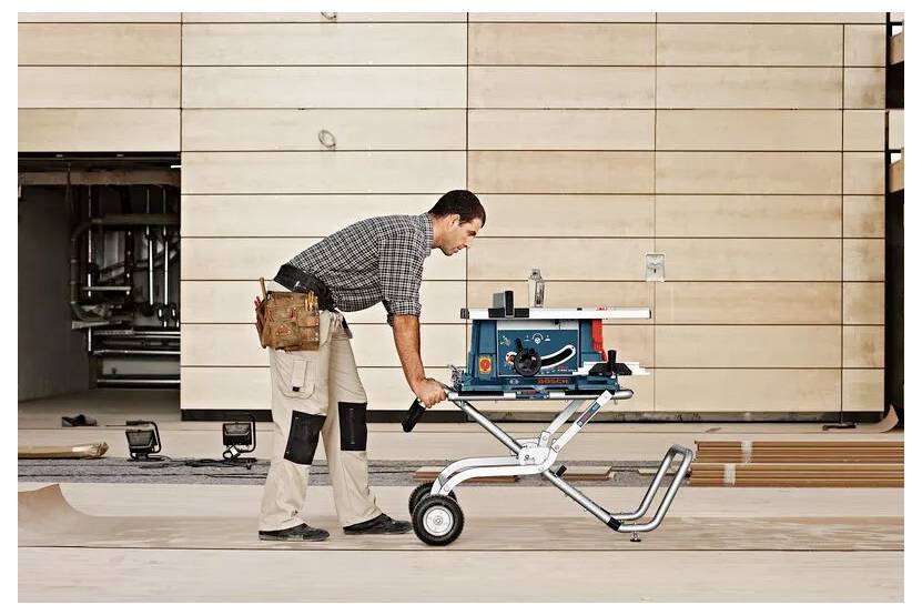 Ein Mann in Arbeitskleidung schiebt eine blaue tragbare Tischkreissäge auf einem fahrbaren Gestell über den Werkstattboden, Werkzeuge sind im Hintergrund sichtbar.