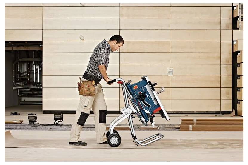 Ein Mann schiebt eine robuste Tischkreissäge auf einem Wagen. Er trägt Arbeitshosen und einen Werkzeuggürtel und geht auf einer Holzplattform in einer Baustelle.