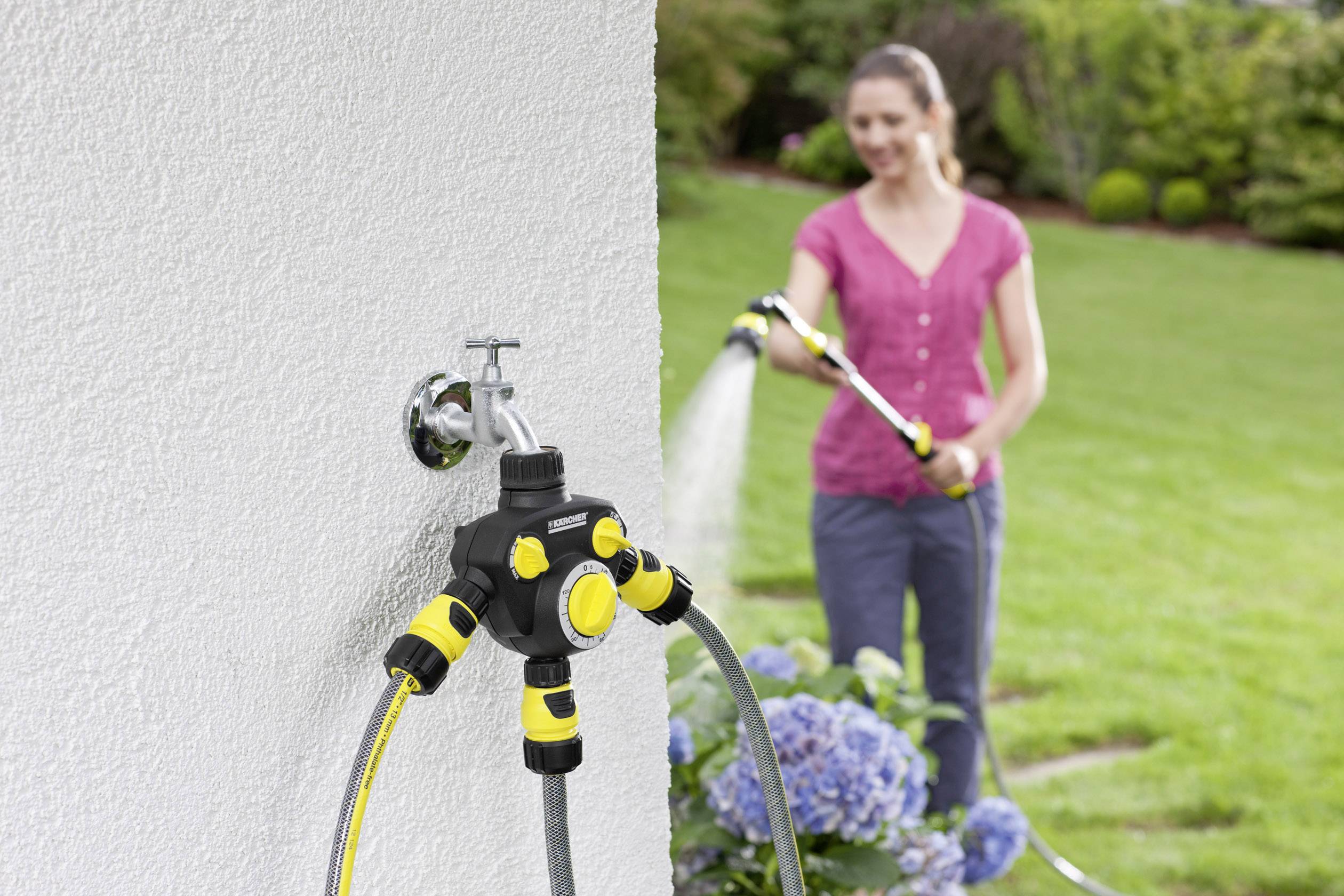 Eine Frau gießt den Garten mit einem Schlauch. Im Vordergrund ist ein an der Wand montierter Wasserverteiler mit mehreren Anschlüssen zu sehen.