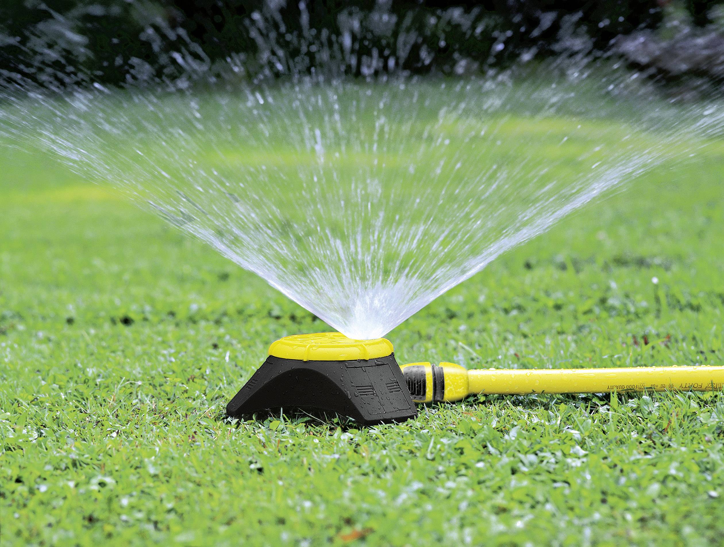 Ein gelber Rasensprenger bewässert grünes Gras im Garten. Wasserstrahlen sprühen in alle Richtungen, um die Fläche gleichmäßig zu bedecken.