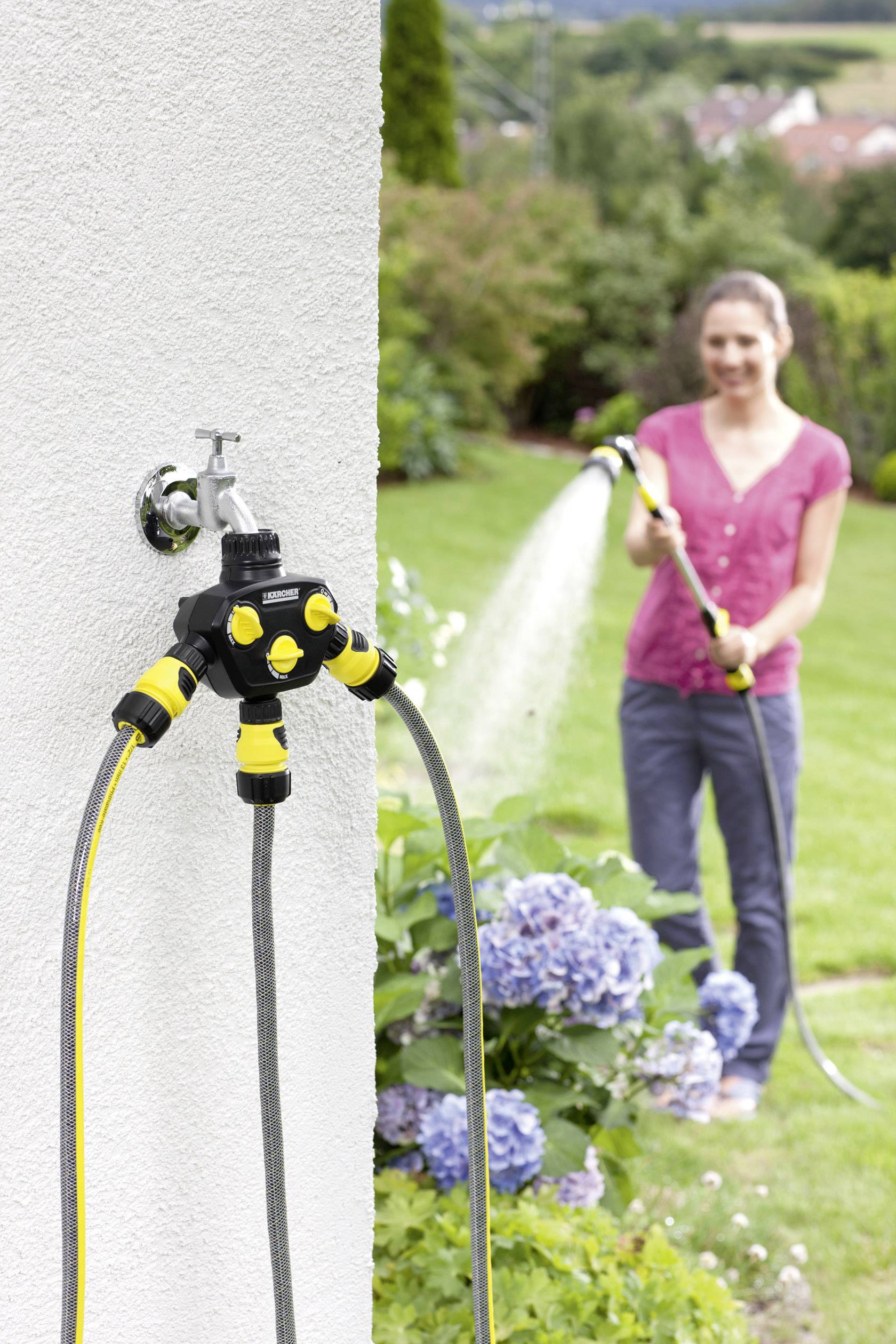 Eine Frau gießt den Rasen mit einem Gartenschlauch. Im Vordergrund ein Wassersprinkler mit mehreren angeschlossenen Schläuchen.