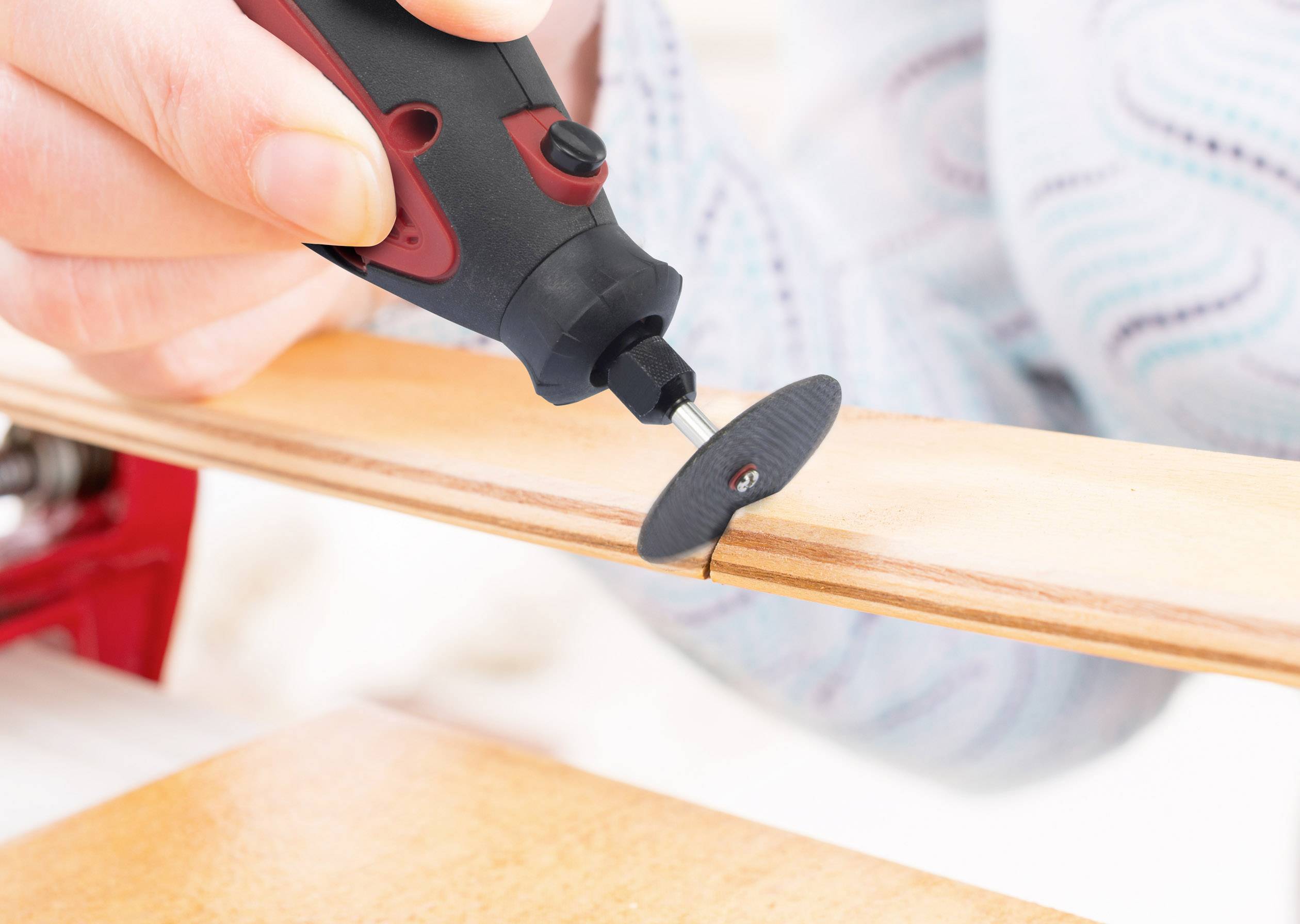 Eine Person nutzt ein rot-schwarzes Dremel-Werkzeug, um präzise in Holz zu schneiden. Die Nahaufnahme zeigt den Schneidevorgang.